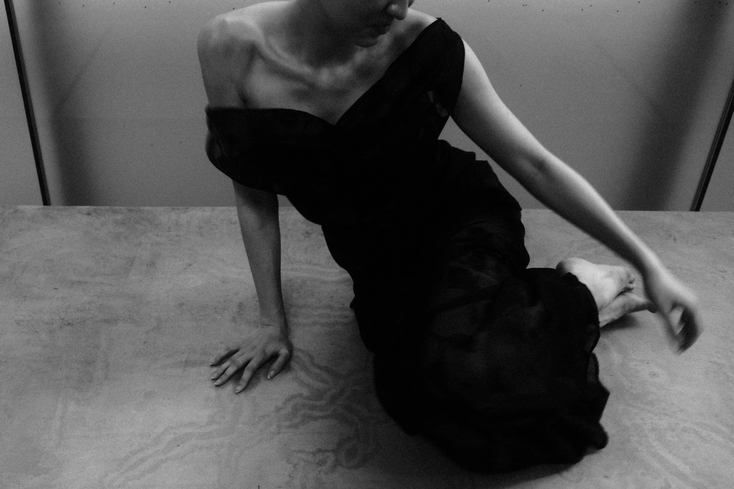 A person in a black dress sitting on a table, leaning on one hand, with the other hand reaching out, against a plain backdrop.