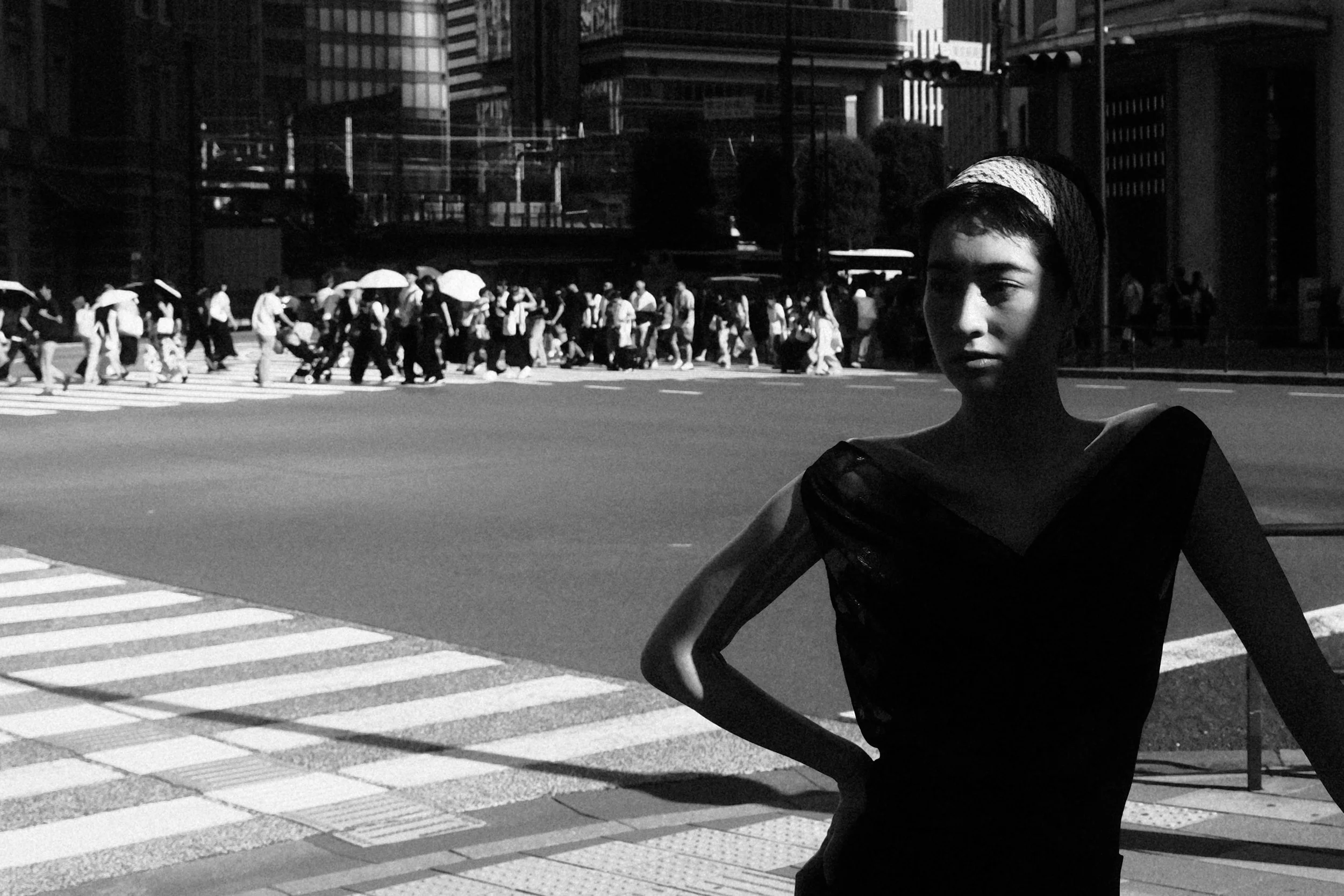 A woman standing on a city crosswalk in shadow, with a busy crowd of pedestrians crossing the street behind her.