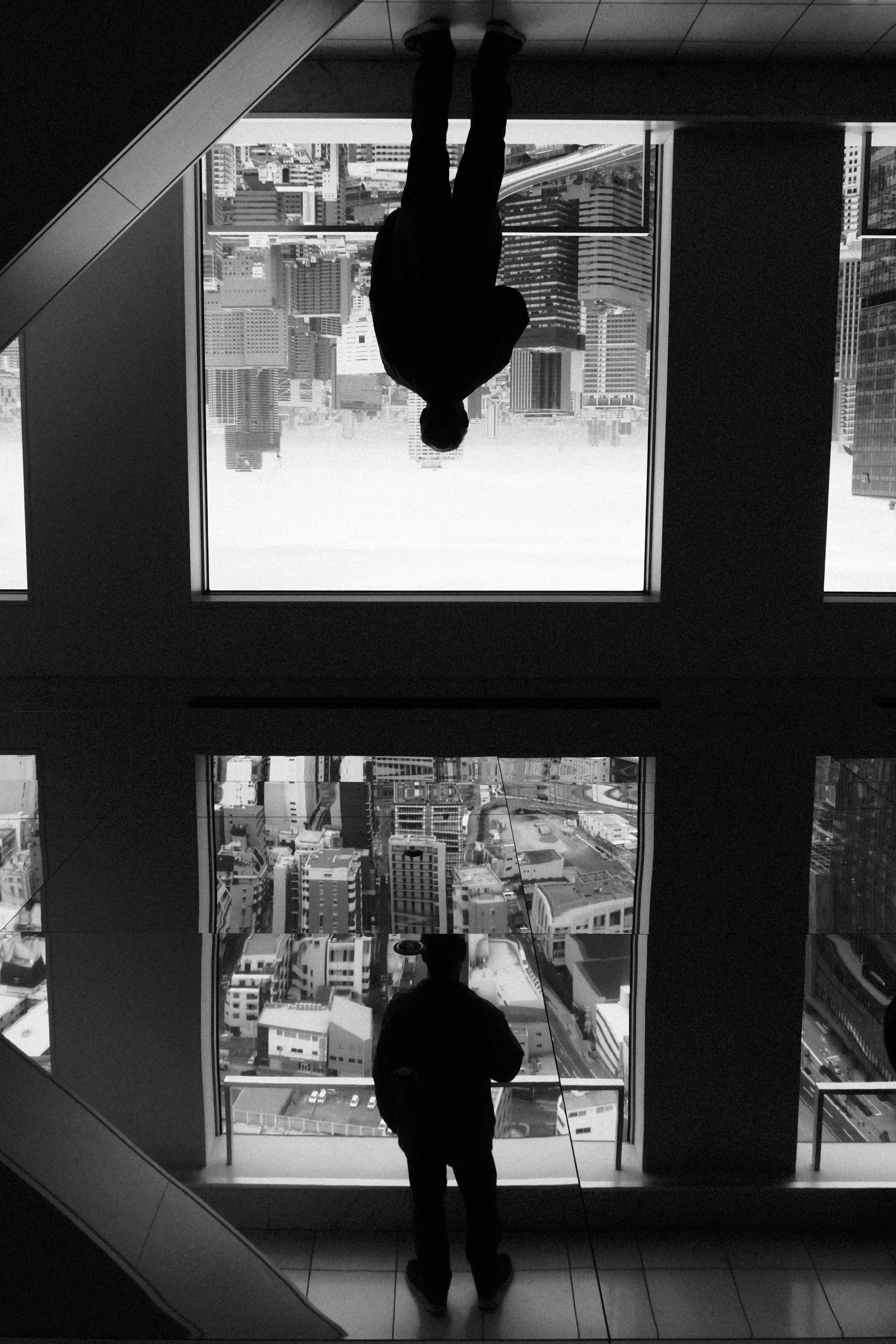Black and white photo of two people inside a tall building, one looking out a large window at a cityscape, the other taking a photo of the view, with the city buildings and streets in the background.