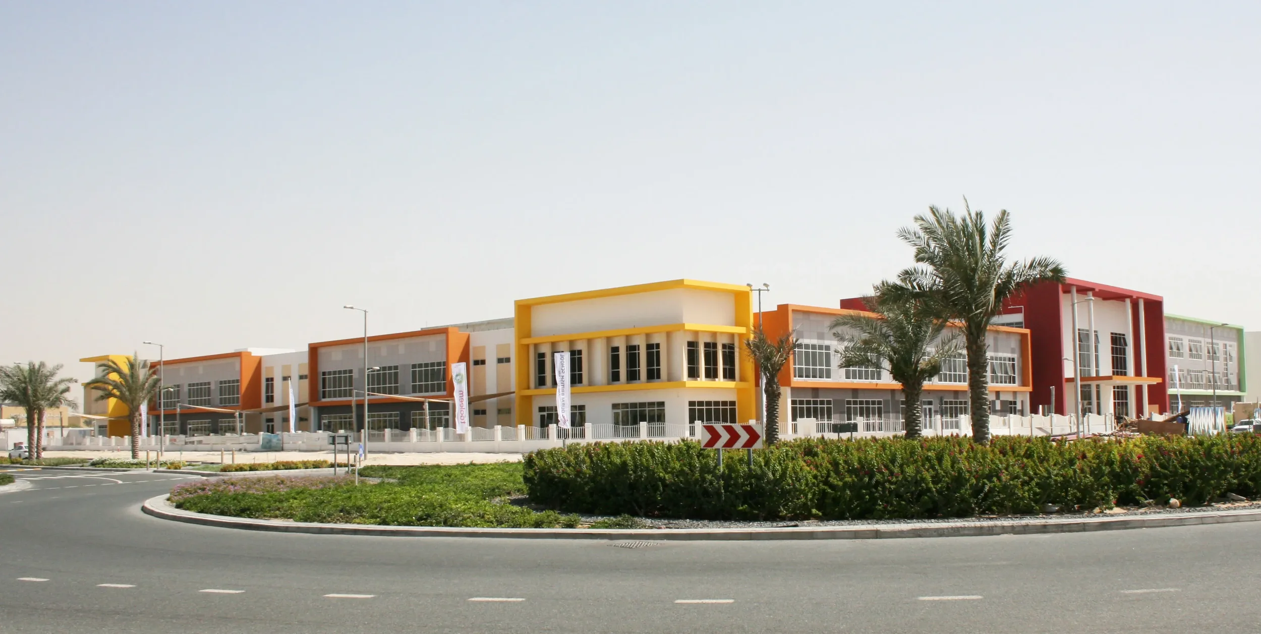 Colorful modern building with large glass windows, surrounded by palm trees and landscaped greenery, situated along a curved road.