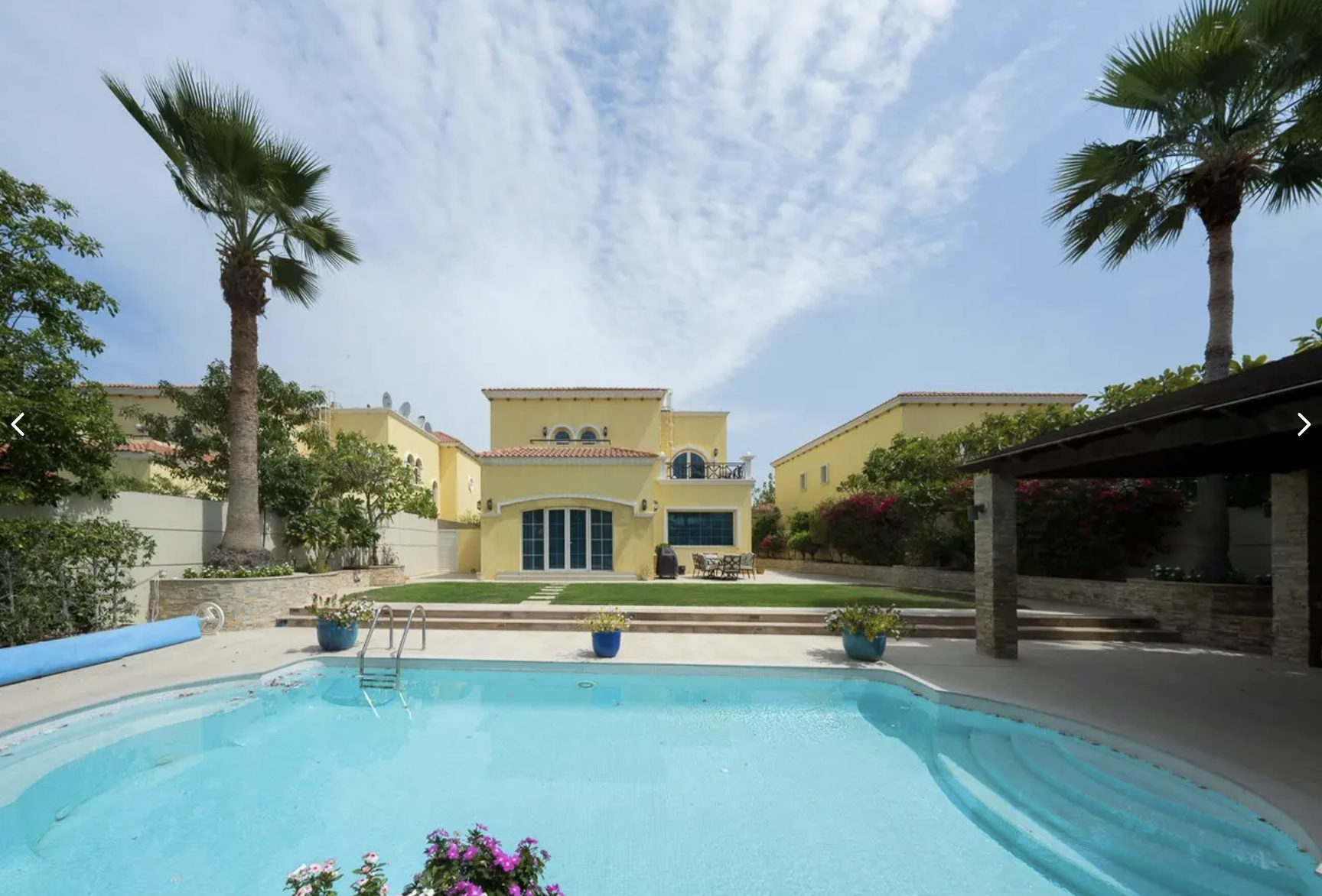 A backyard with a swimming pool, two palm trees, and a yellow house with a patio and stairs, under a partly cloudy sky.