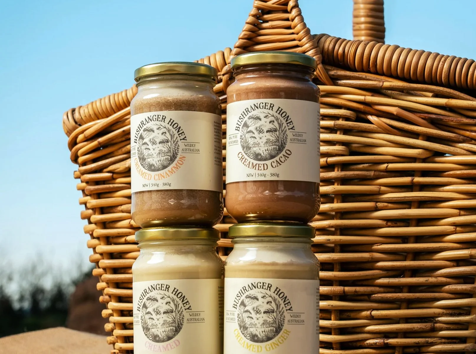 Four jars of Bushranger Honey products, including creamed cinnamon, creamed cacao, creamed ginger, and creamed honey, arranged on a wicker picnic basket outdoors with a clear sky background.