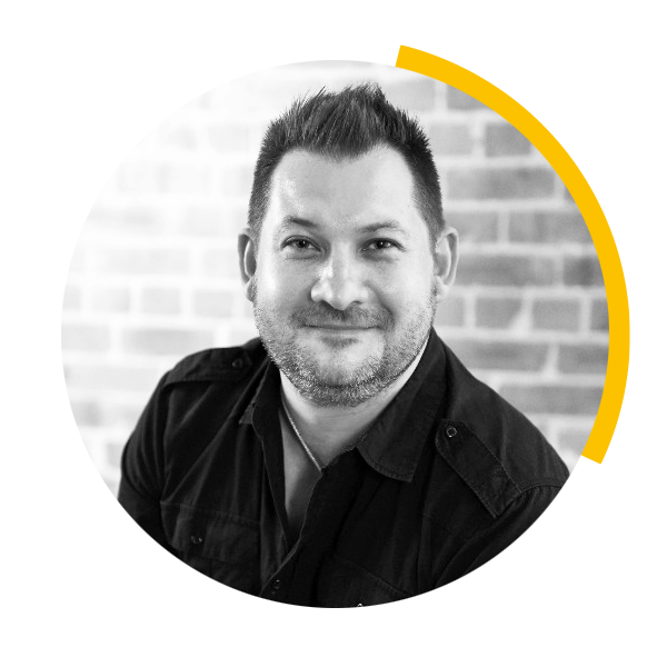 Black and white portrait of a man with short dark hair, slight beard, smiling, wearing a dark shirt, sitting in front of a brick wall.