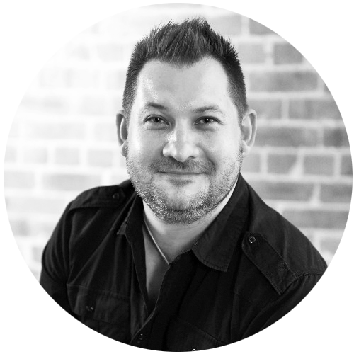 Black and white portrait of a smiling man with short dark hair and facial hair, wearing a dark shirt, sitting against a brick wall background.