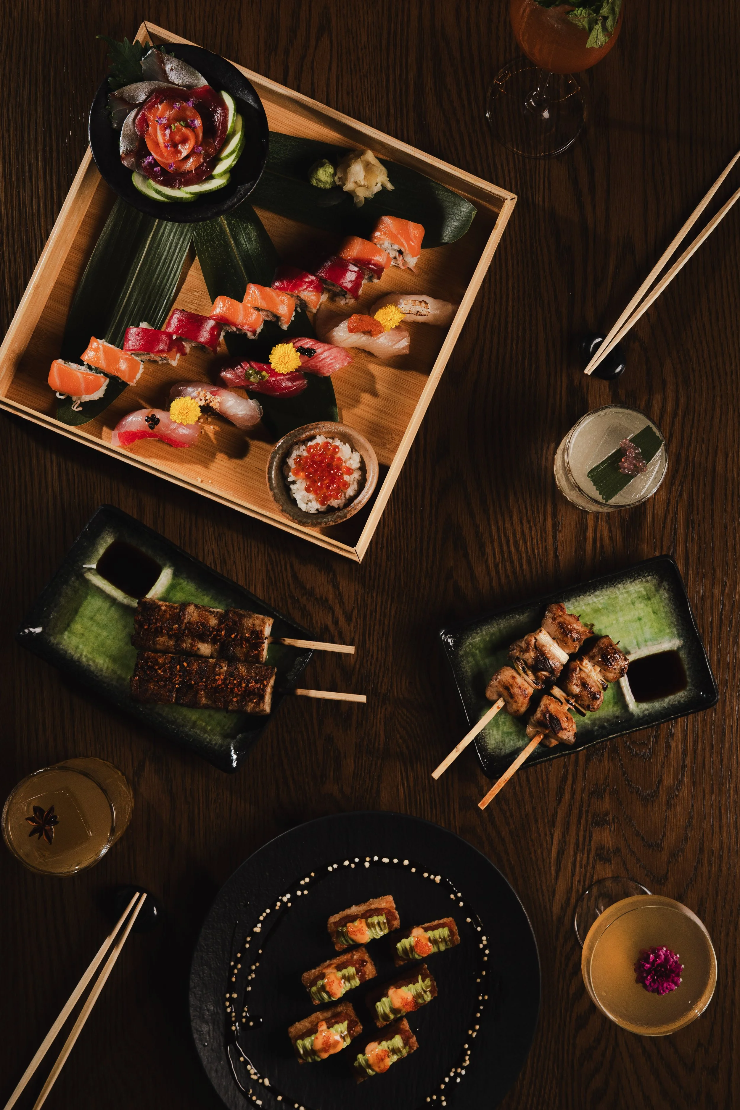 Various types of sushi on black and wooden plates, garnished with flowers and accompanied by drinks, chopsticks, and soy sauce on a dark wooden table.