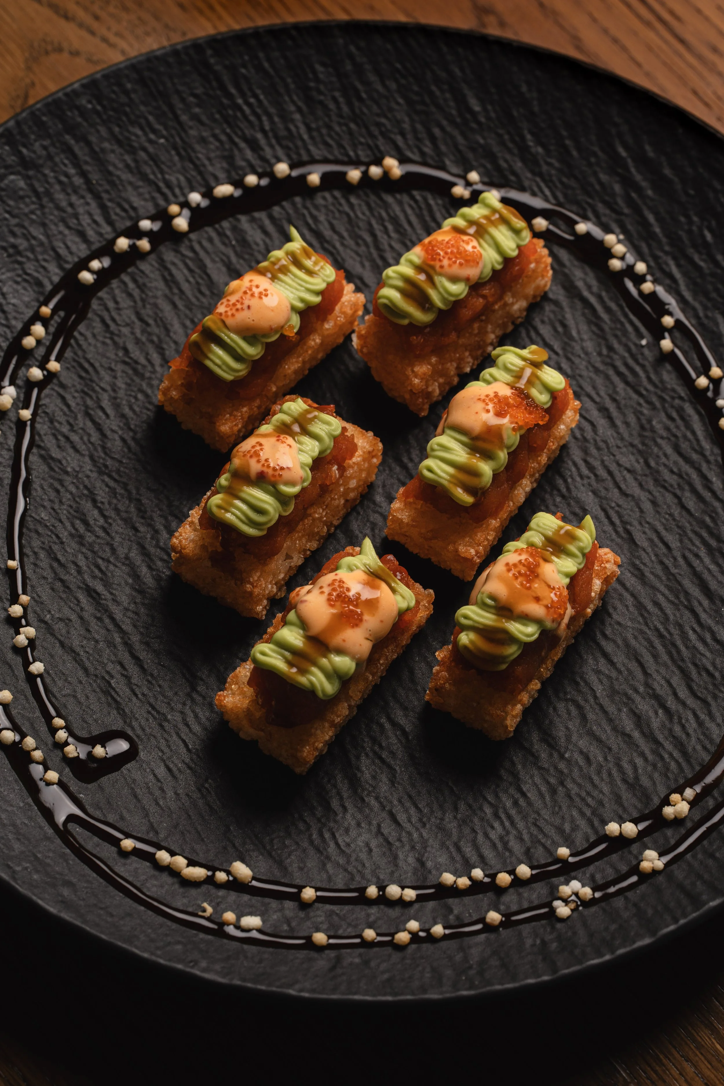Six pieces of sushi with eel, green sauce, and orange roe on top, arranged on a black plate with decorative sauce and crunch around the edges.