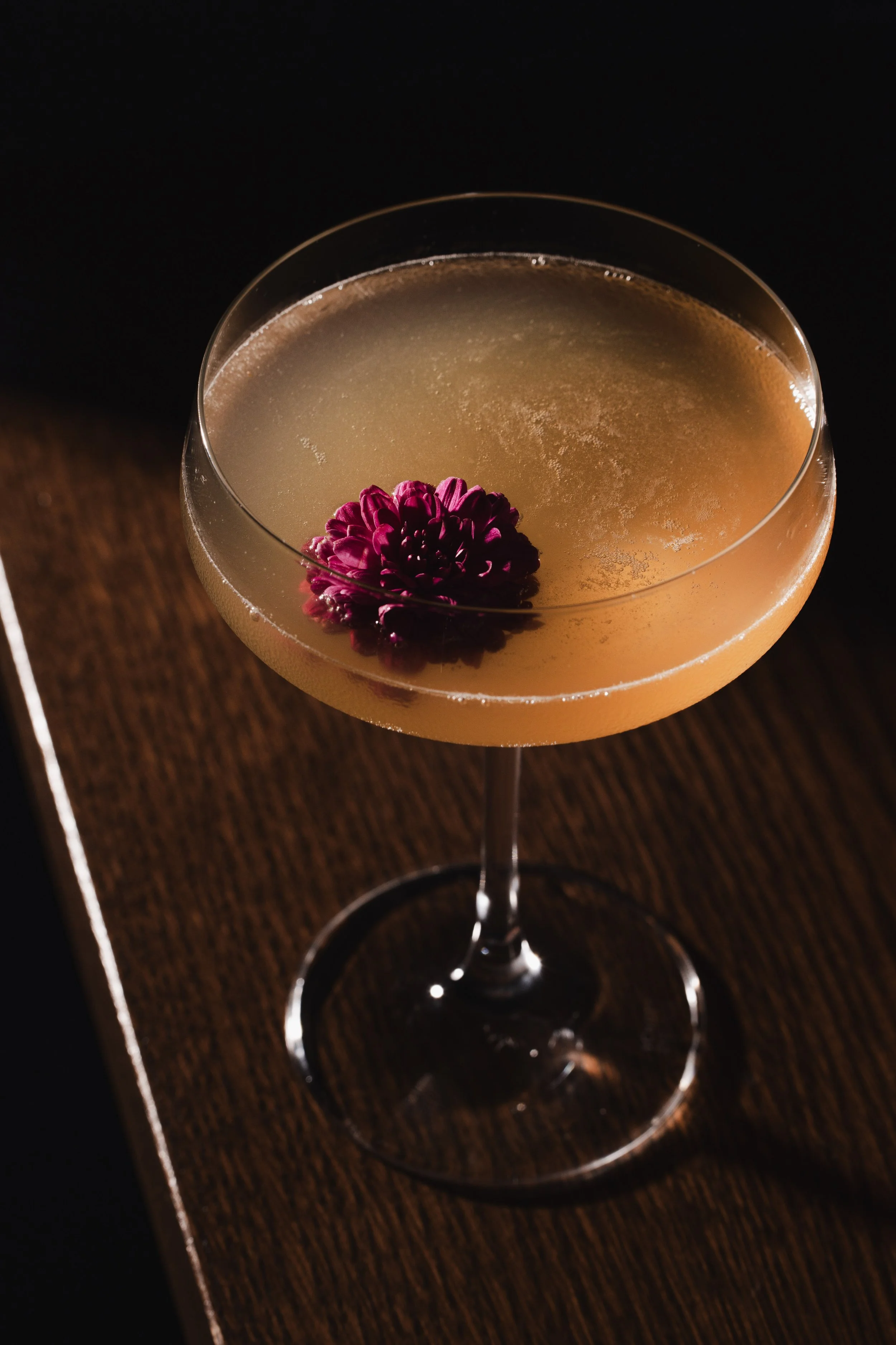 A cocktail glass filled with a light amber-colored drink, garnished with a small cluster of pinkish flowers. The glass is on a dark wooden surface against a black background.