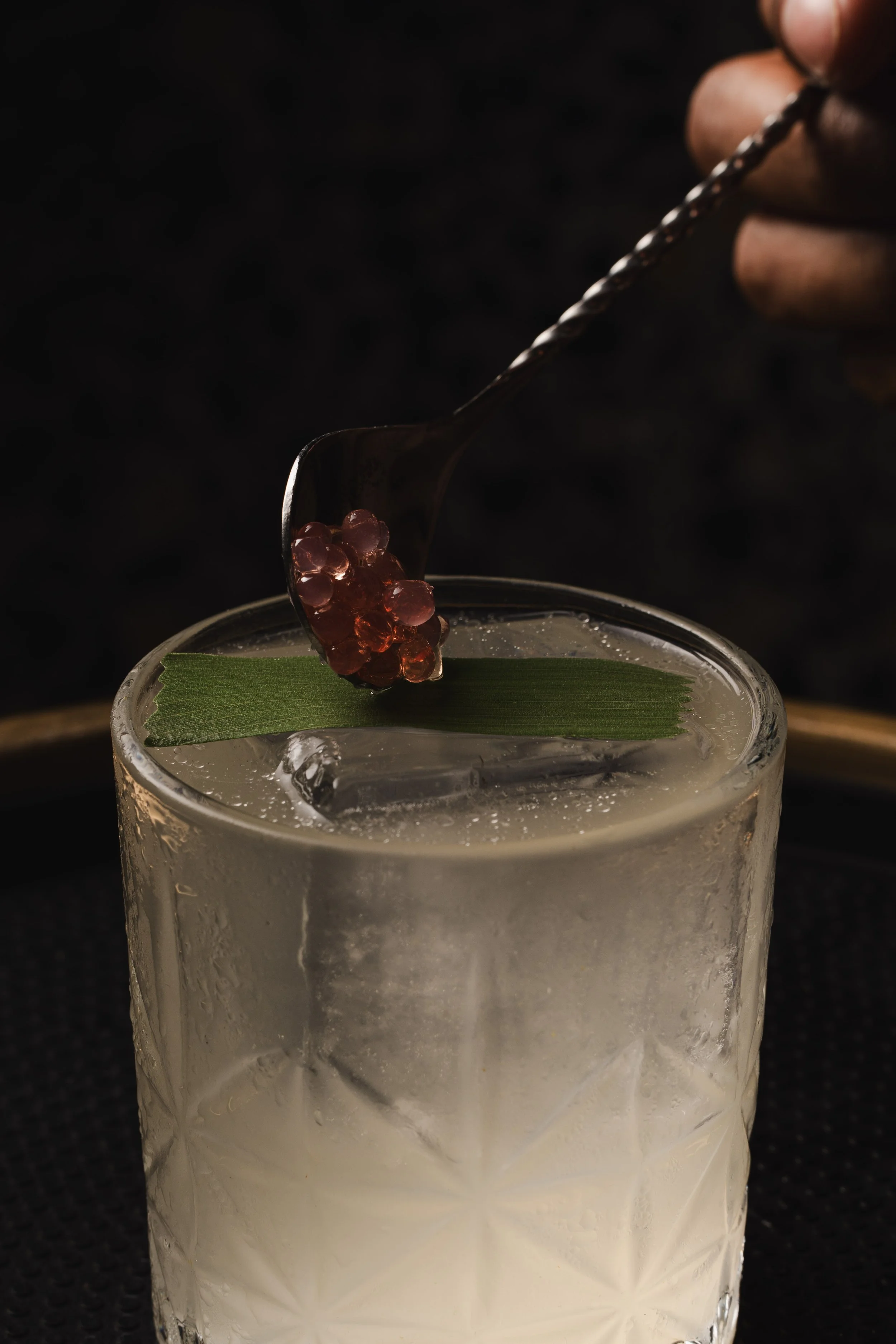 Close-up of a hand holding a twisted spoon with small pinkish-red pearls above a frosted glass filled with a white creamy liquid. There is a green leaf on the rim of the glass, against a dark background.