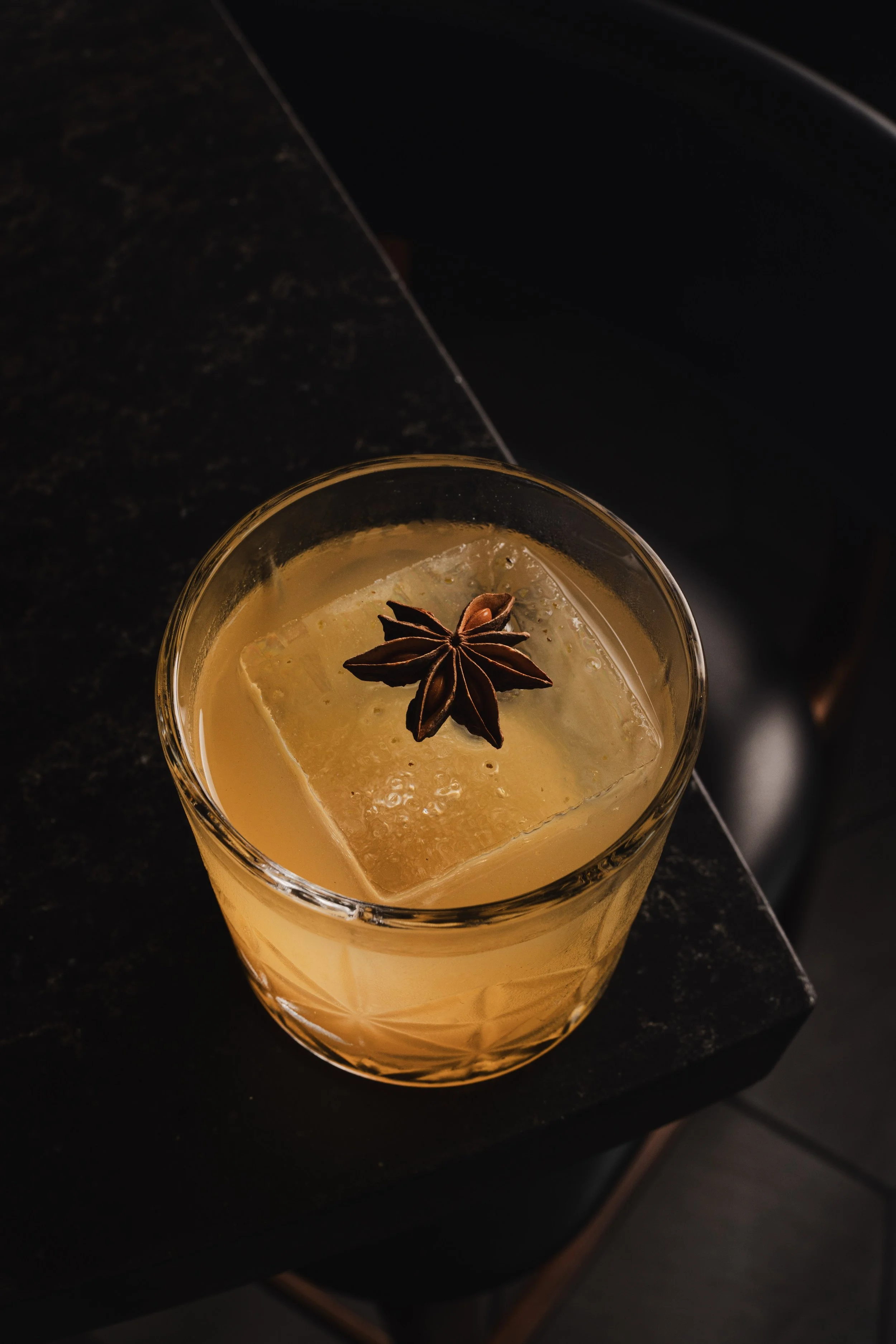 Close-up of a glass of whiskey with ice and a star anise garnish on top, placed on a dark surface.