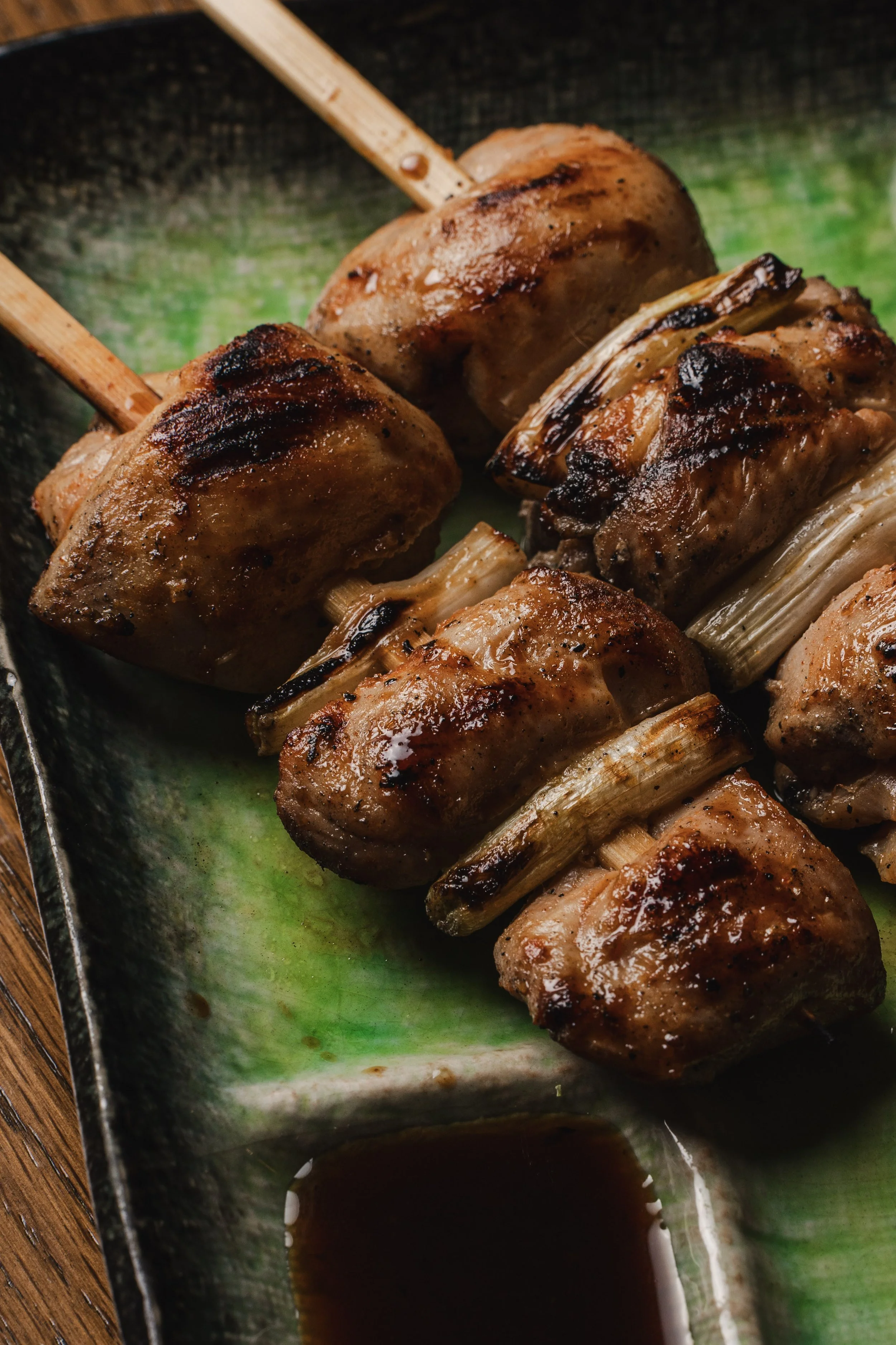 Close-up of grilled skewered meat with charred grill marks, placed on a green glazed ceramic dish with soy sauce at the bottom.