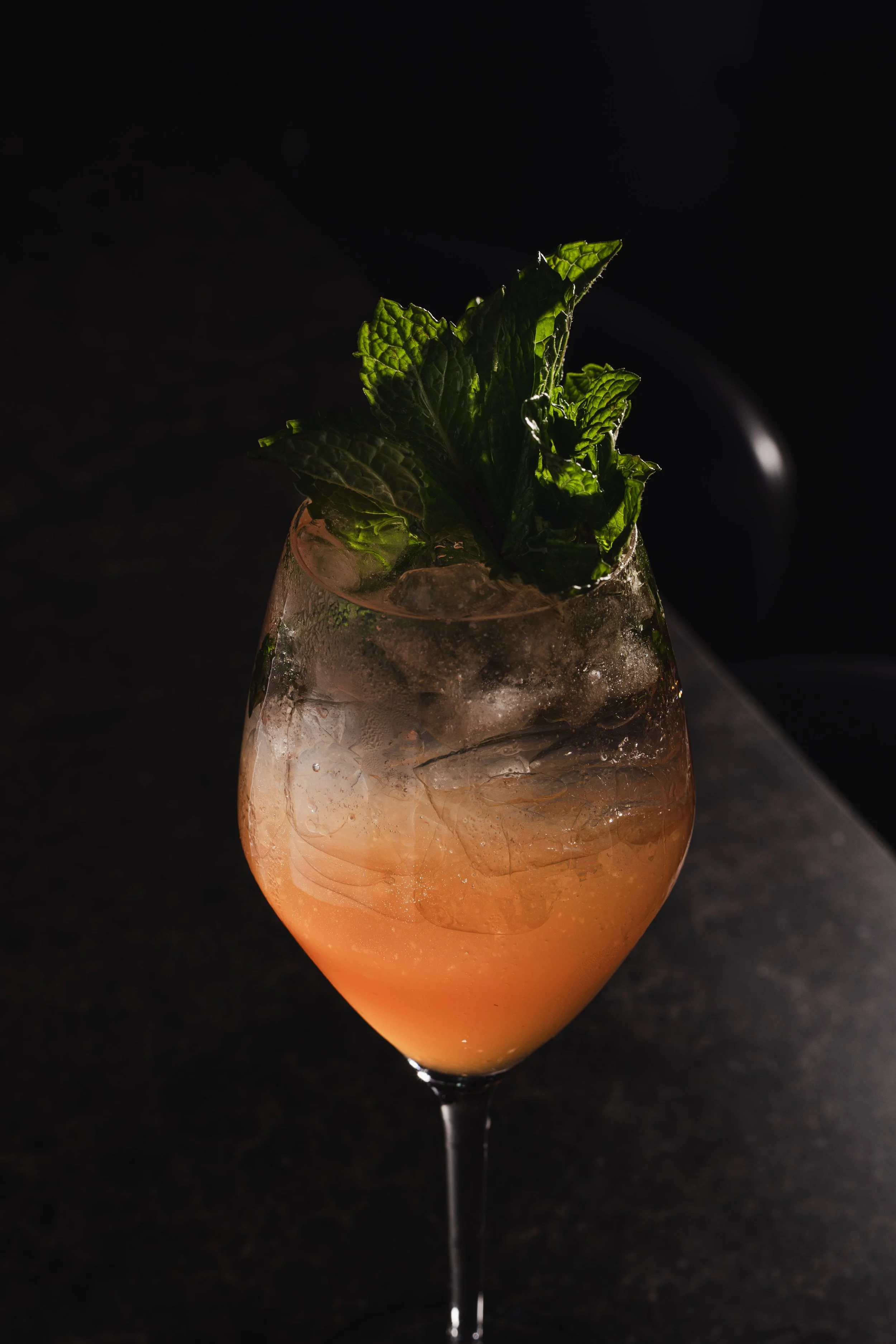 A cocktail in a tall, elegant glass with a gradient of orange and pink hues, topped with fresh green mint leaves, set against a dark background.