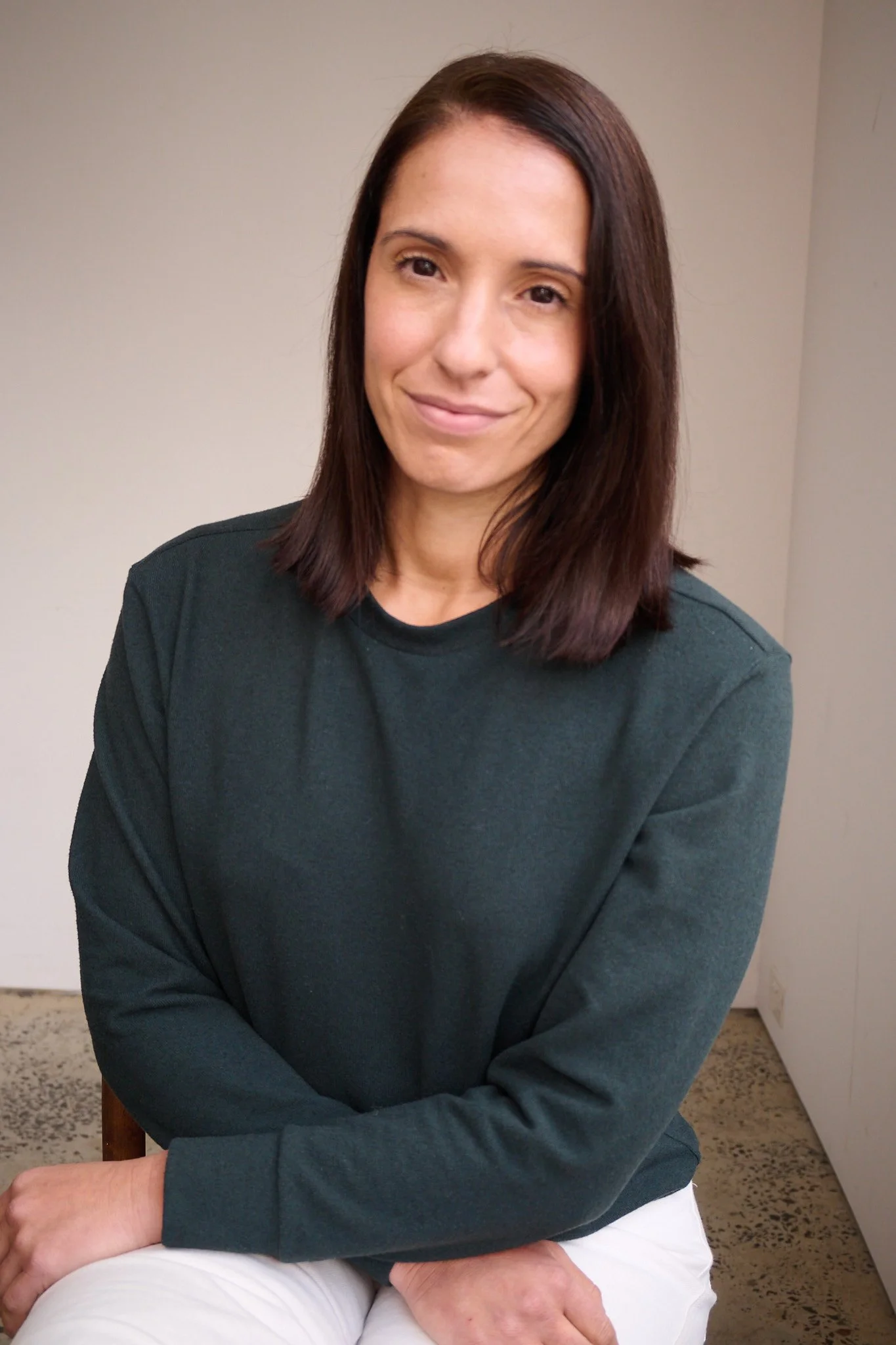 A woman with shoulder-length brown hair, wearing a dark green long-sleeve shirt and white pants, sitting on a chair in front of a plain wall.