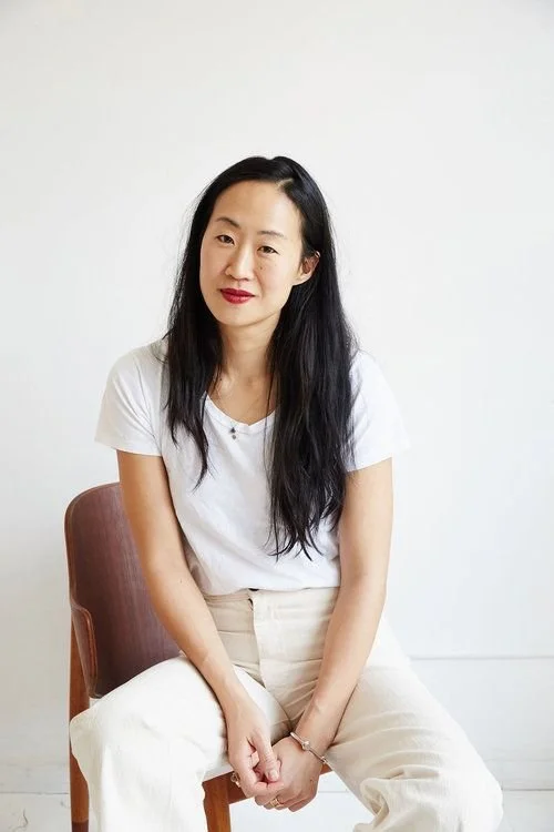 A woman with long black hair and red lipstick sitting on a pink chair against a plain white wall, wearing a white t-shirt and beige pants.