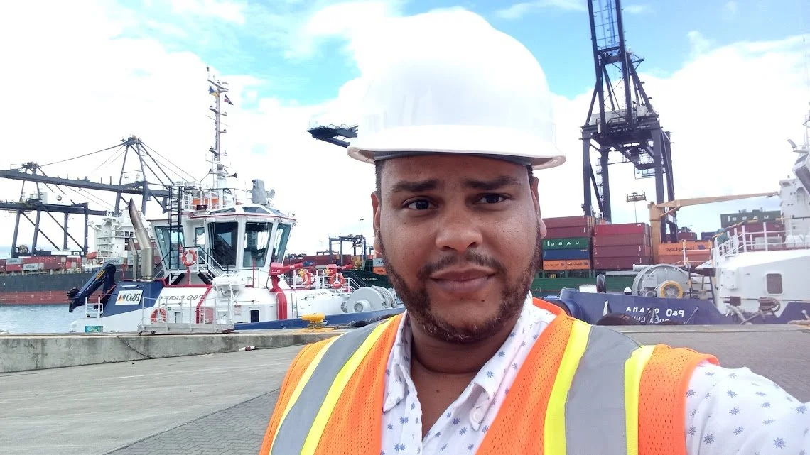 Hombre con casco de seguridad y chaleco reflectante en puerto marítimo, con barcos y grúas de carga en el fondo.