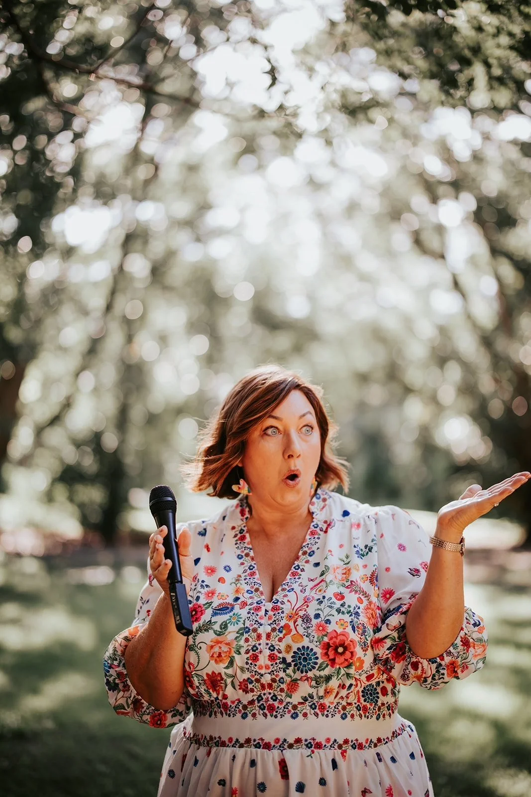 Krista Webb, Marriage Celebrant is holding a microphone outdoors, with an expressive look and gesture, surrounded by blurred trees and sunlight.