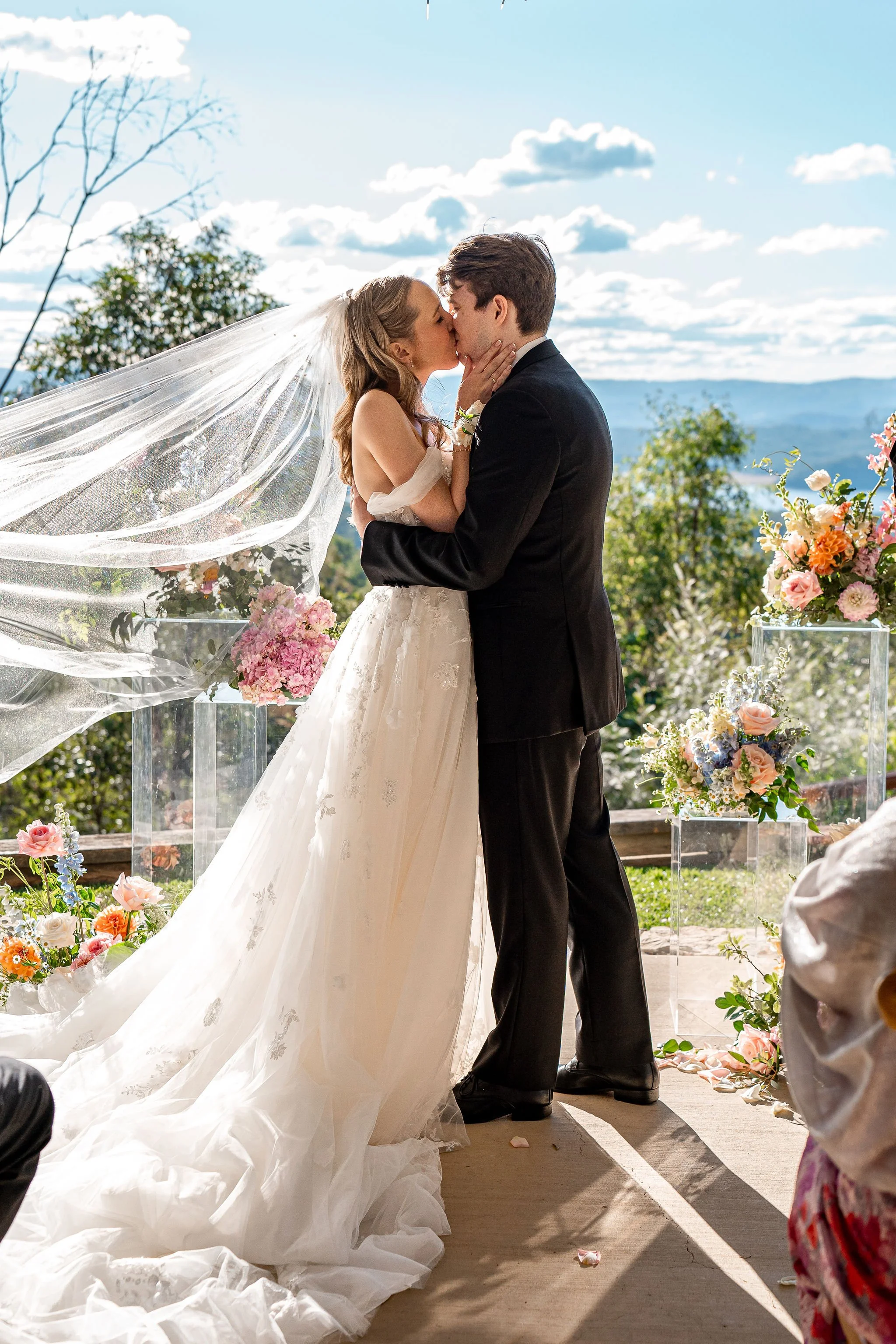 A bride and groom kissing during their outdoor wedding ceremony with a mountainous landscape and blue sky in the background, decorated with flowers.