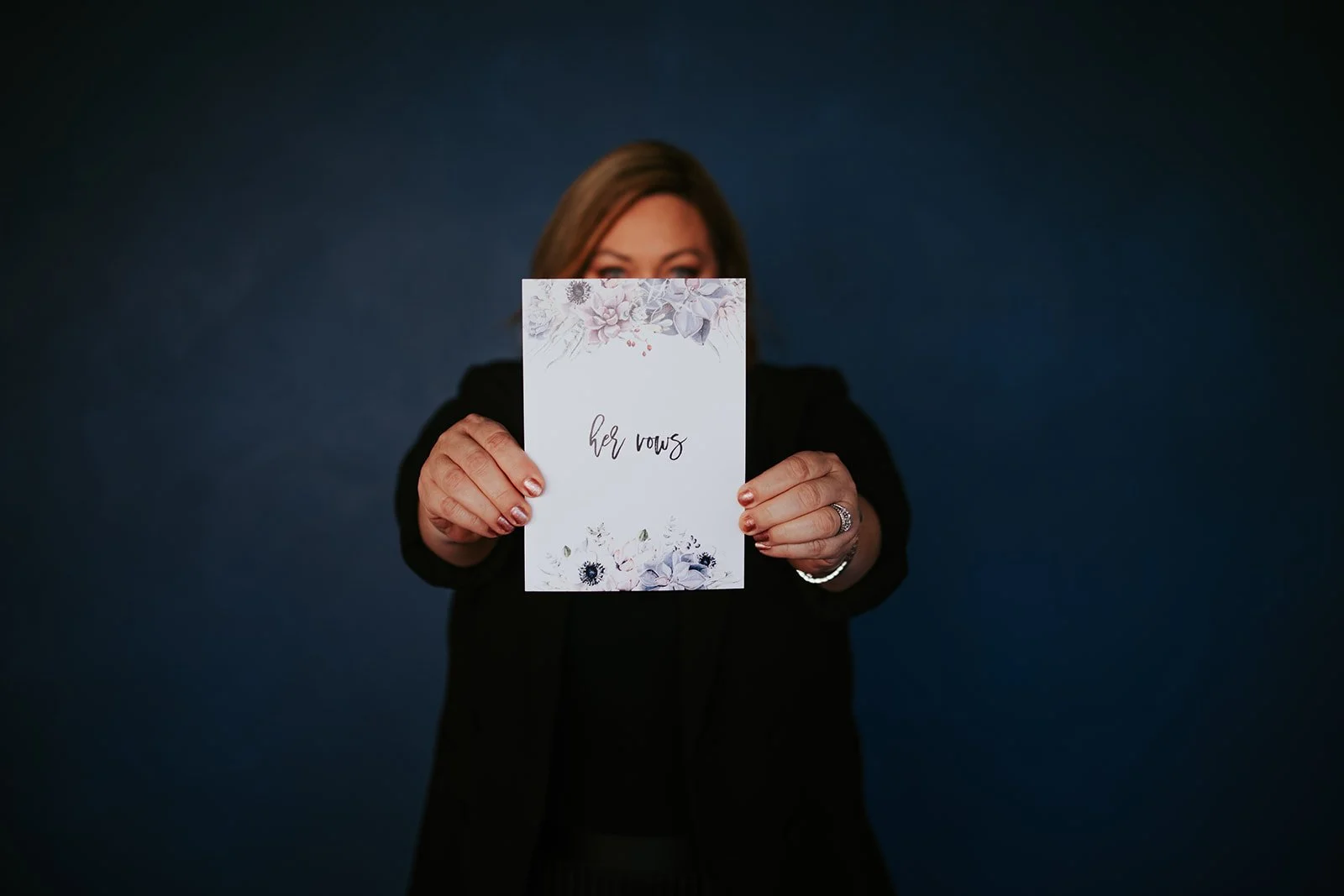 Woman holding a card that says "her vows" with floral design on a dark blue background