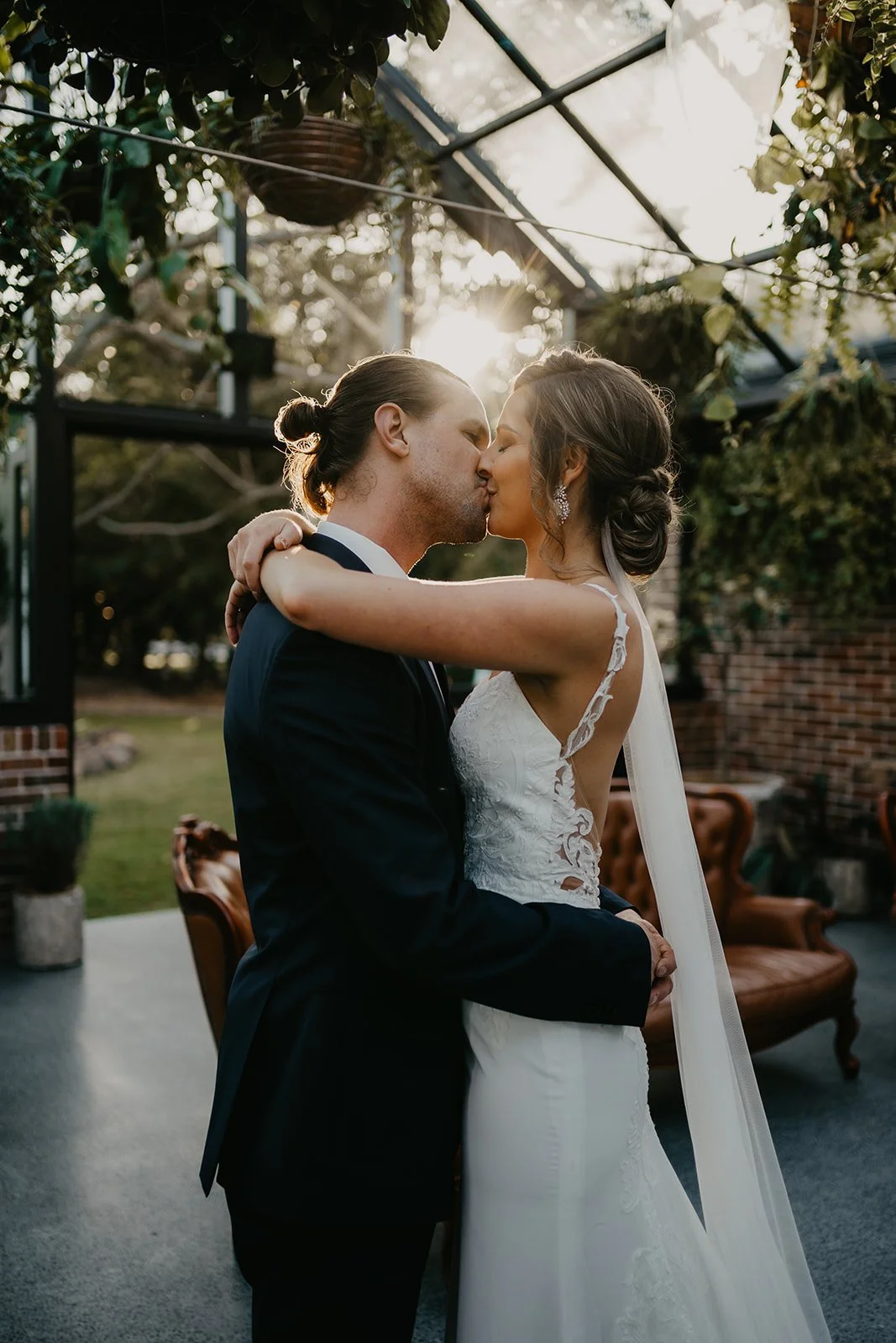 A bride and groom kissing during their wedding ceremony outdoors, with sunlight shining through trees in the background.