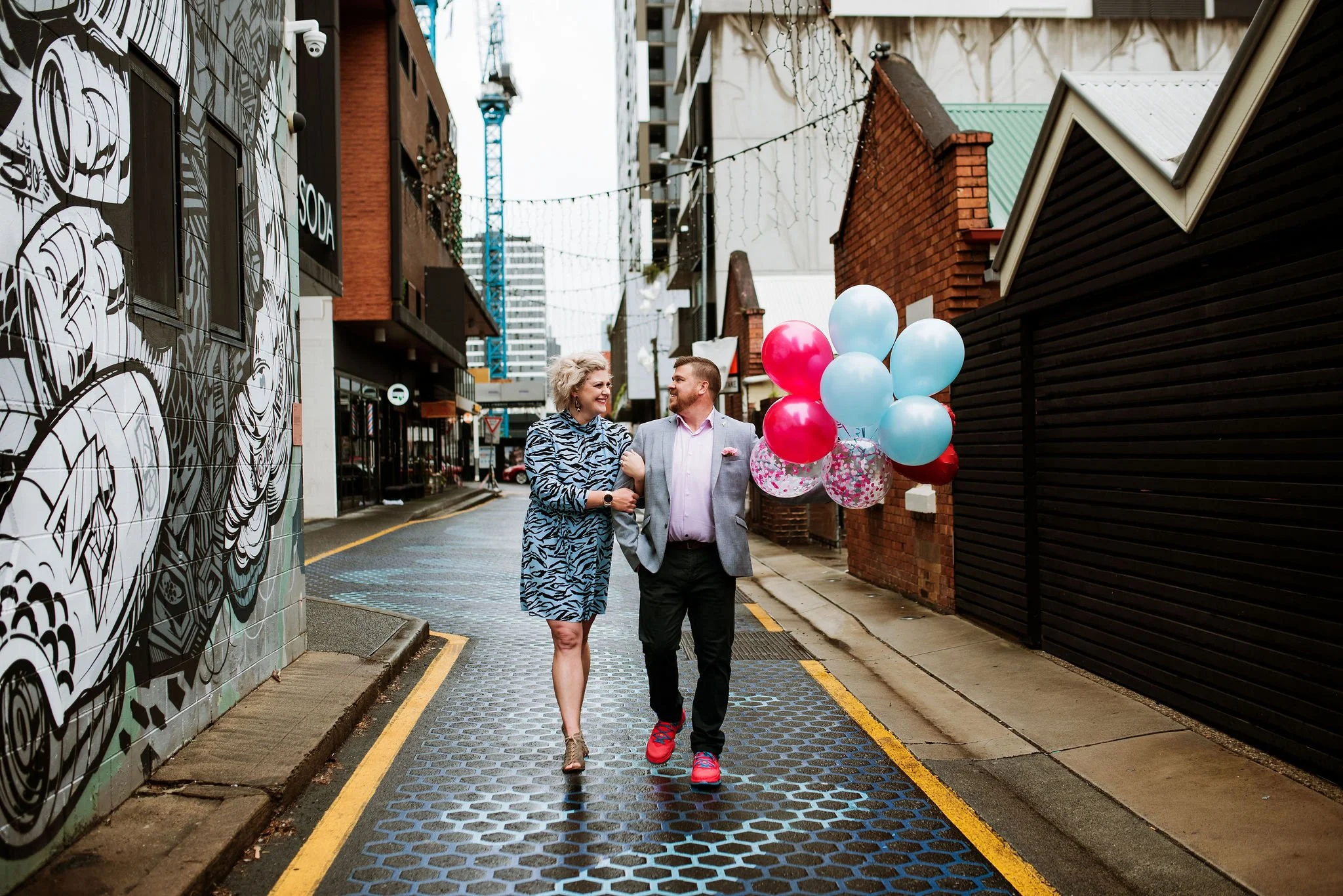 A couple walking arm in arm down a city alley with a bunch of pink and blue balloons, smiling at each other.