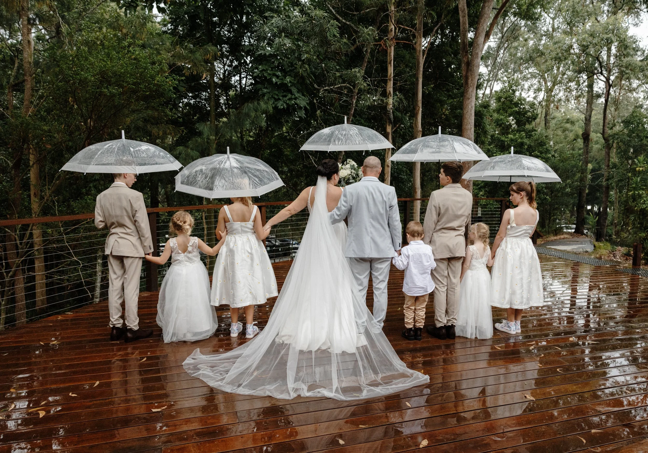 A wedding party standing on a wet wooden deck outdoors with raindrops falling, holding umbrellas, surrounded by trees.