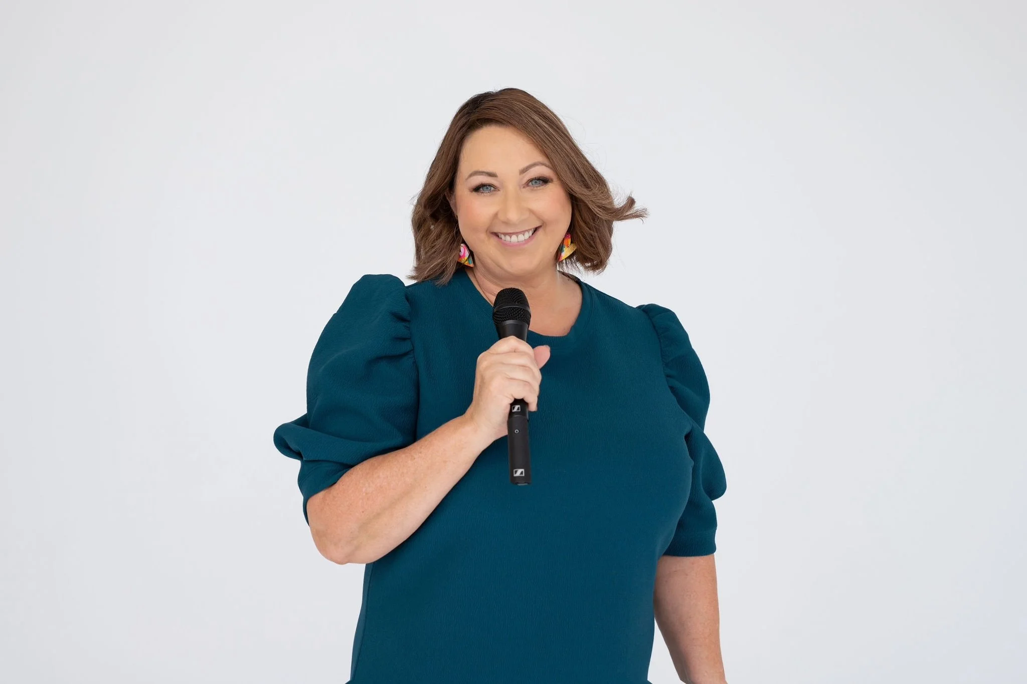 Celebrant Krista Webb from Celebrated Moments, with shoulder-length brown hair, wearing a teal dress with puffed sleeves, holding a microphone and smiling at the camera against a plain white background.