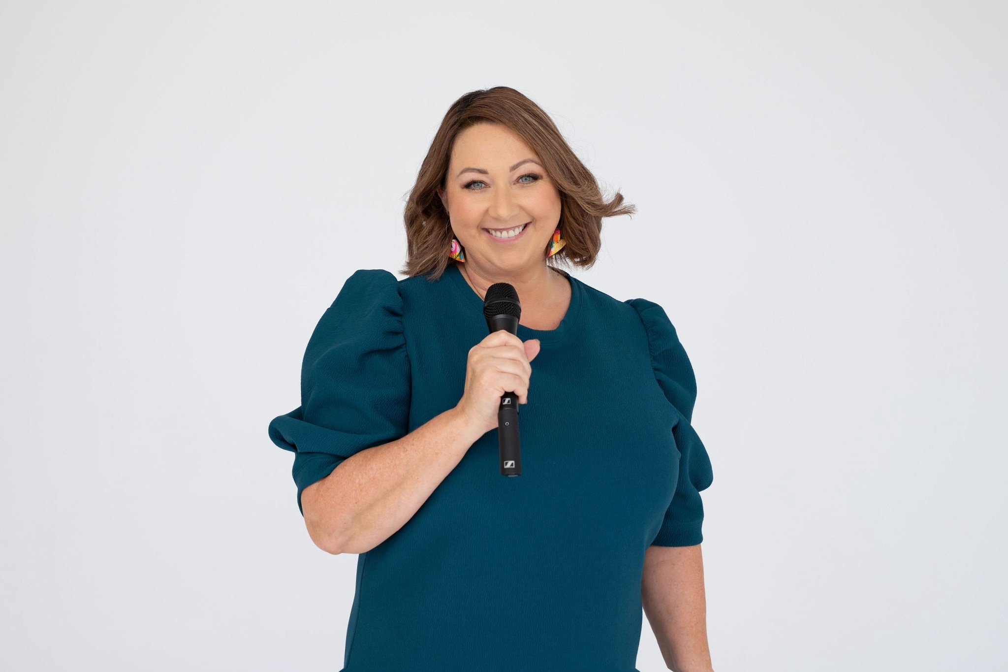 Celebrant Krista Webb from Celebrated Moments, with shoulder-length brown hair, wearing a teal dress with puffed sleeves, holding a microphone and smiling at the camera against a plain white background.