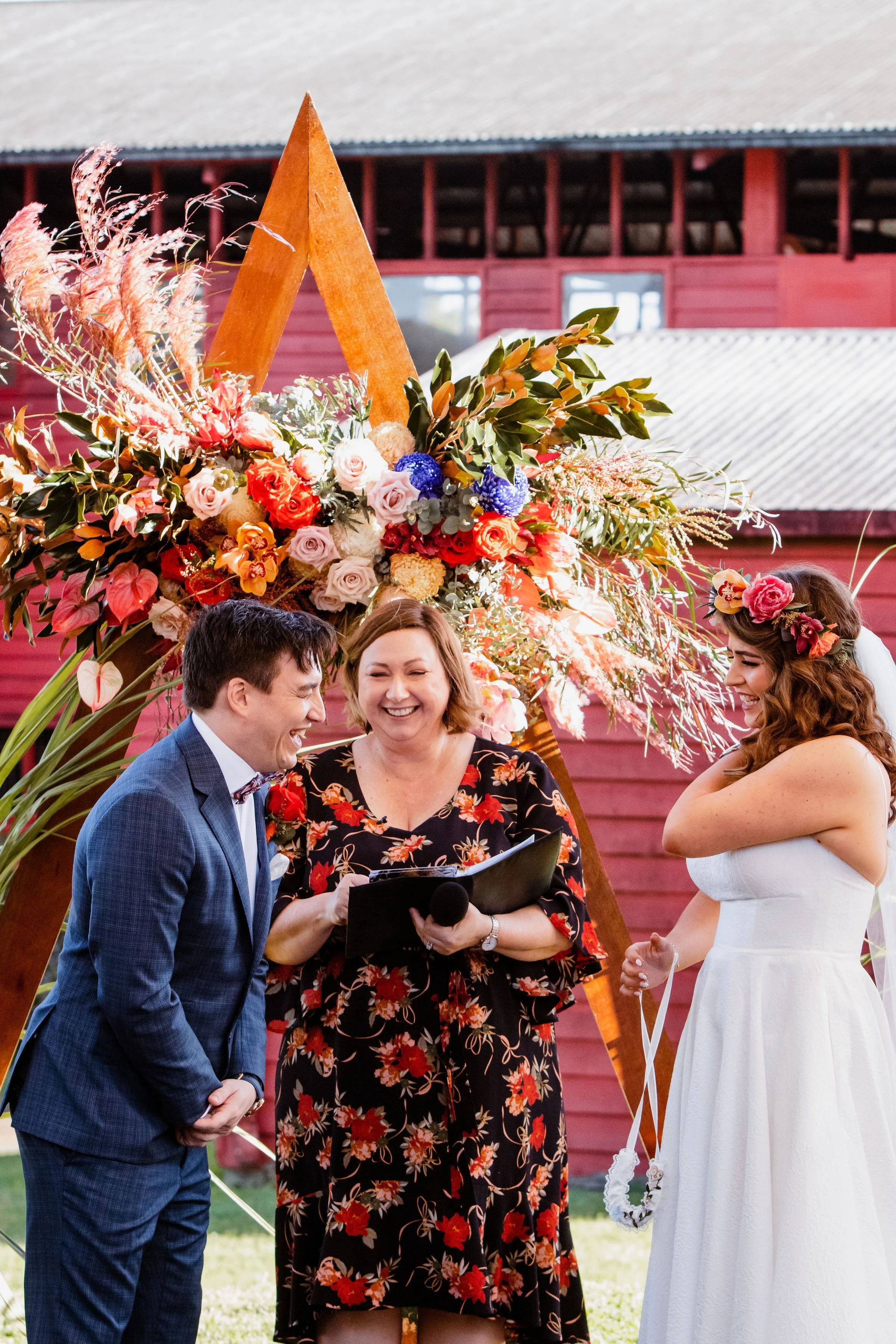 A wedding ceremony with a couple exchanging vows, standing in front of a floral arch with an officiant laughing, outside a red barn.
