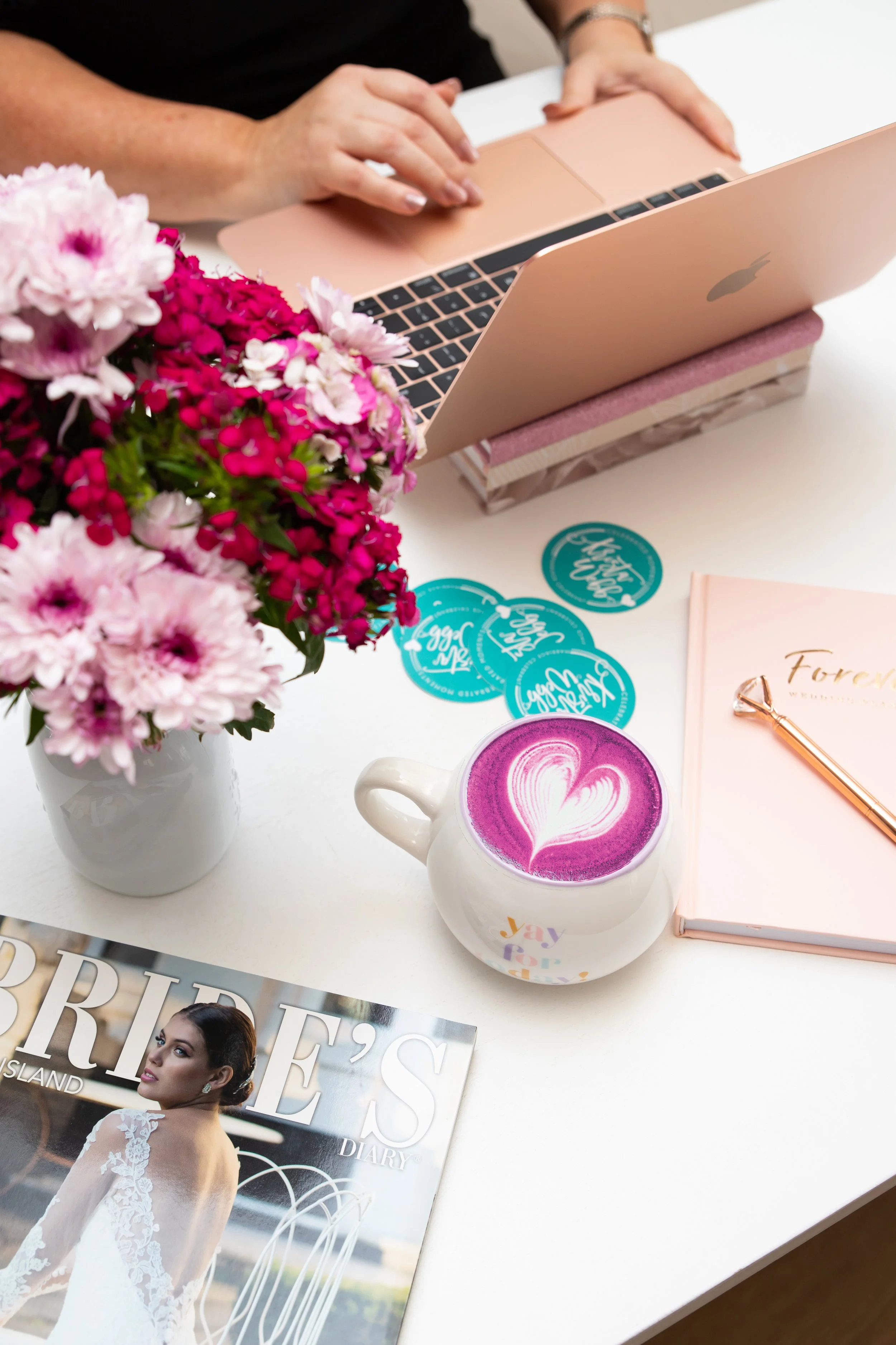 A desk with pink flowers in a white vase, a pink latte with a heart foam art, a notebook labeled 'Forever', a magazine titled 'Bride's Diary', some teal-colored coasters, and a person working on a pink laptop.