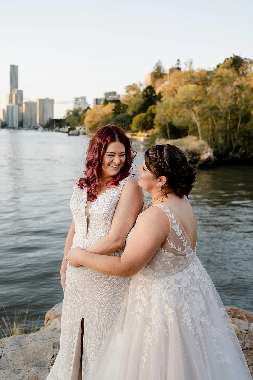Two women in wedding dresses sharing a joyful moment by the water with city buildings and trees in the background.