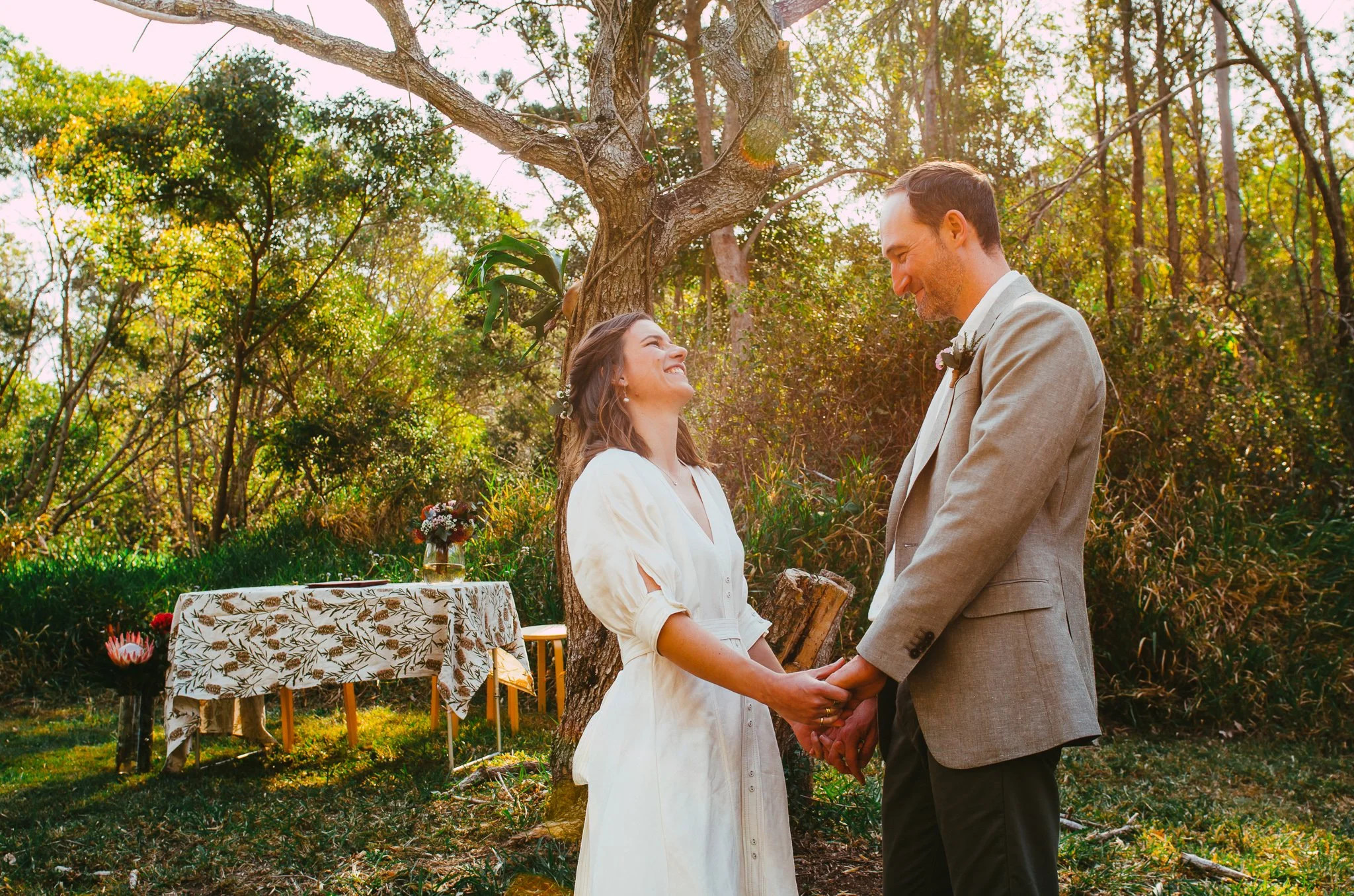 A bride and groom holding hands and smiling at each other during an outdoor wedding ceremony under a tree, with sunlight shining through the branches, and a decorated table with flowers in the background.