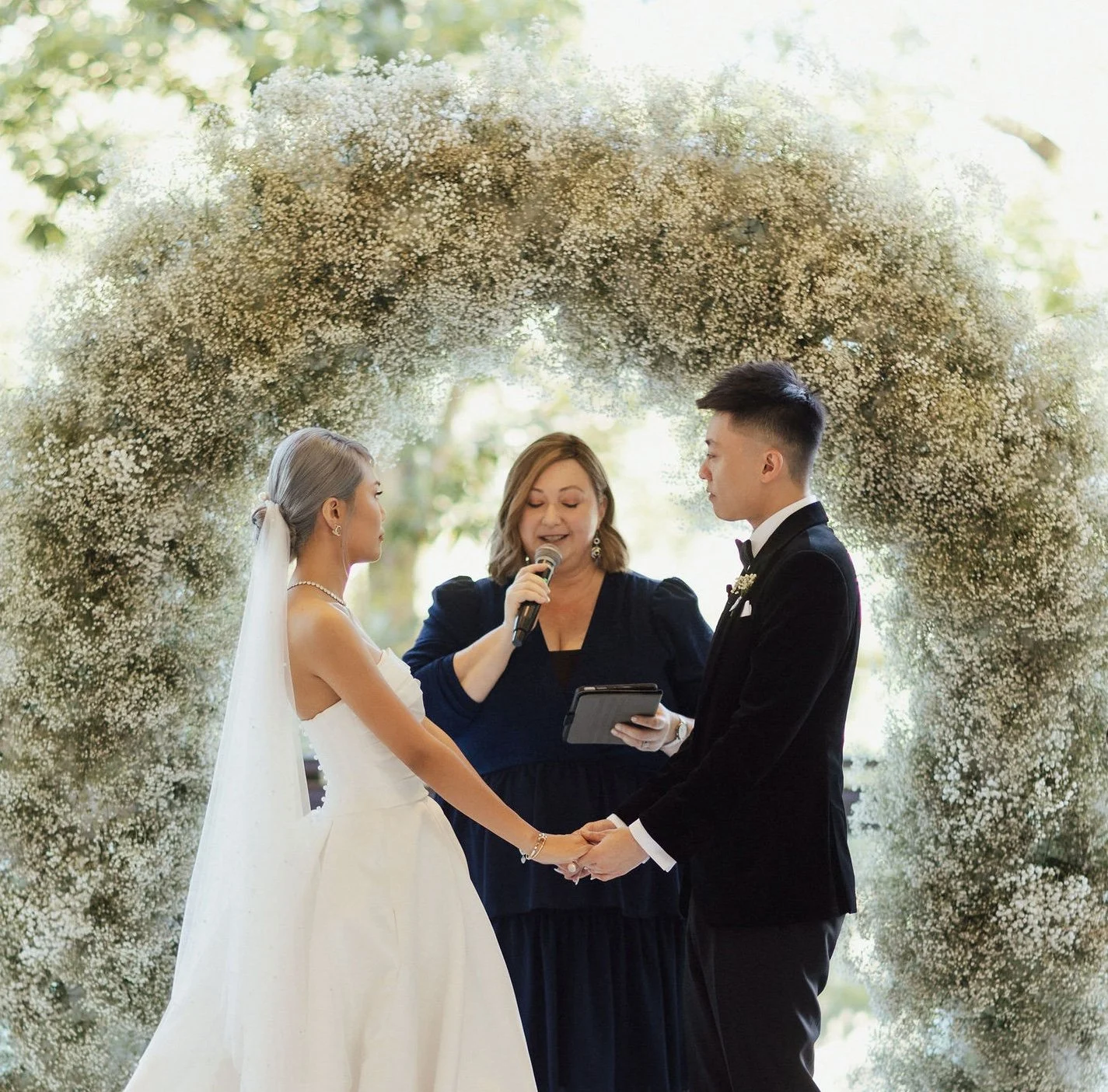 A bride and groom holding hands during their wedding ceremony, standing in front of a floral archway with Marriage Celebrant, Krista Webb, reading vows.