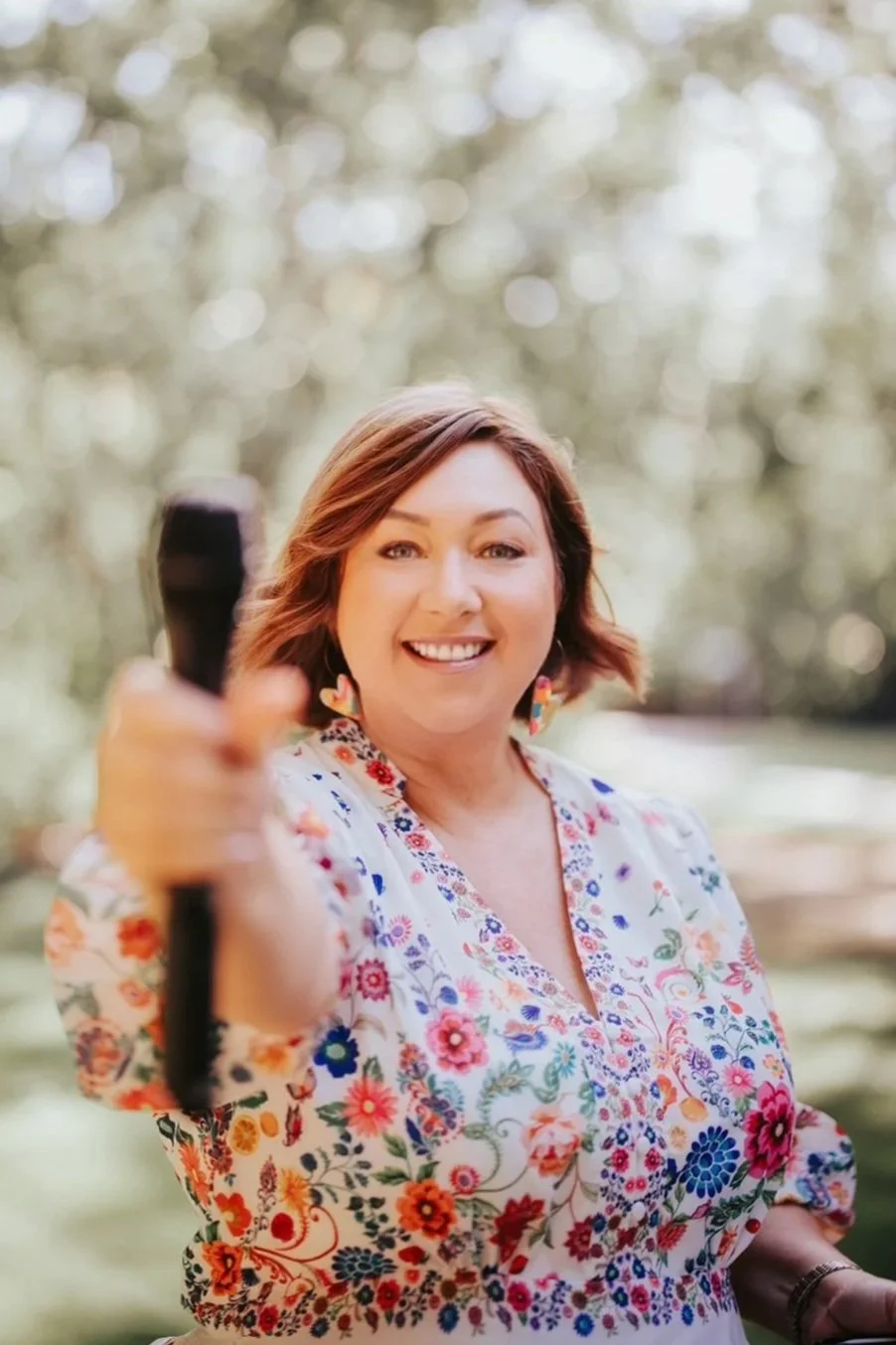 Krista Webb, Marriage Celebrant, smiles and points towards the camera against a blurred green outdoor background.