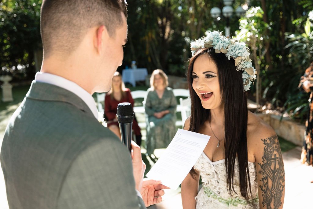 A Bride with a flower crown and tattoos smiling during her outdoor wedding ceremony as a groom reads vows.