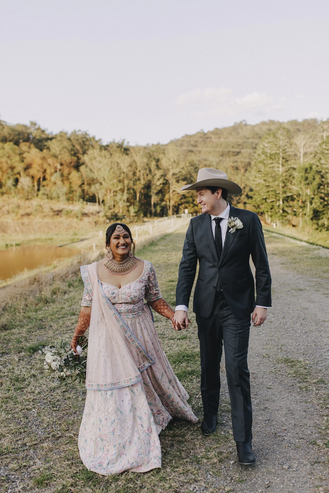A newlywed couple walking hand in hand outdoors, smiling at each other. The bride is wearing a traditional pink and silver wedding dress with jewelry, and the groom is in a dark suit with a cowboy hat. The background includes a pond, trees, and a dir