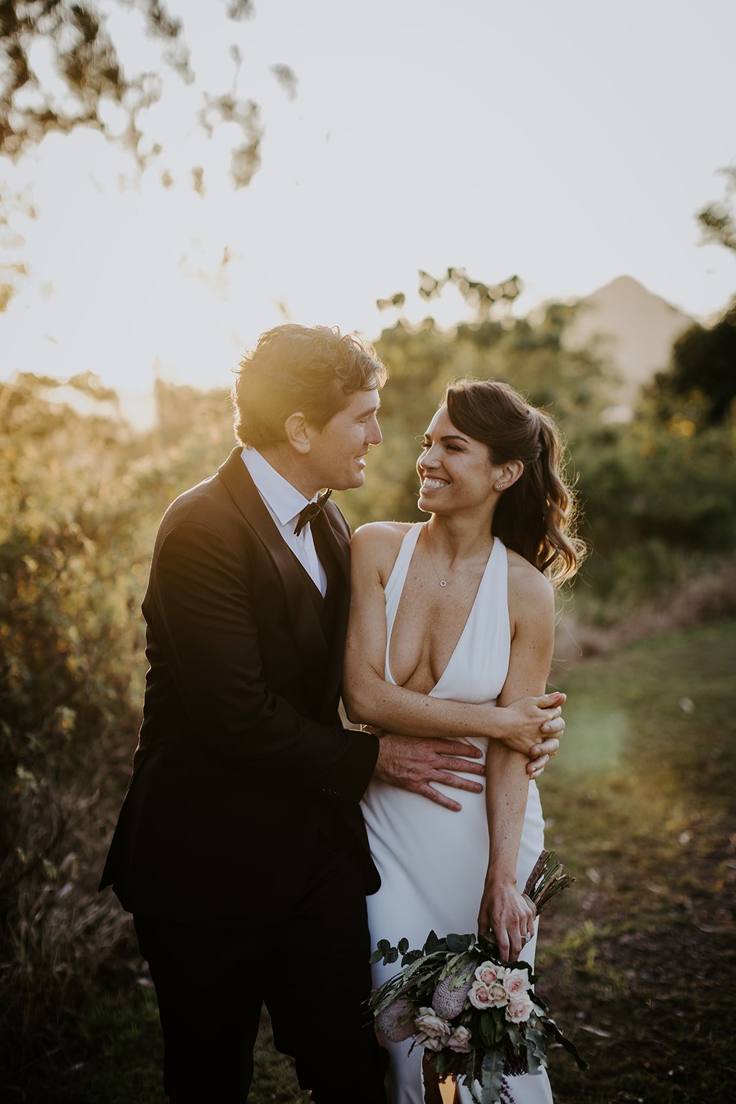A bride and groom standing outdoors during sunset, smiling at each other. The bride is in a white gown holding a bouquet, and the groom is in a black tuxedo.