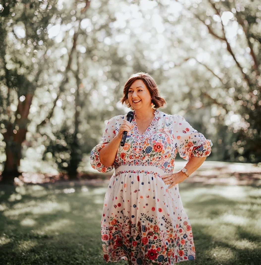 Celebrant Krista Webb from Celebrated Moments in a floral dress holding a microphone, smiling outdoors in a park or garden setting with trees in the background.