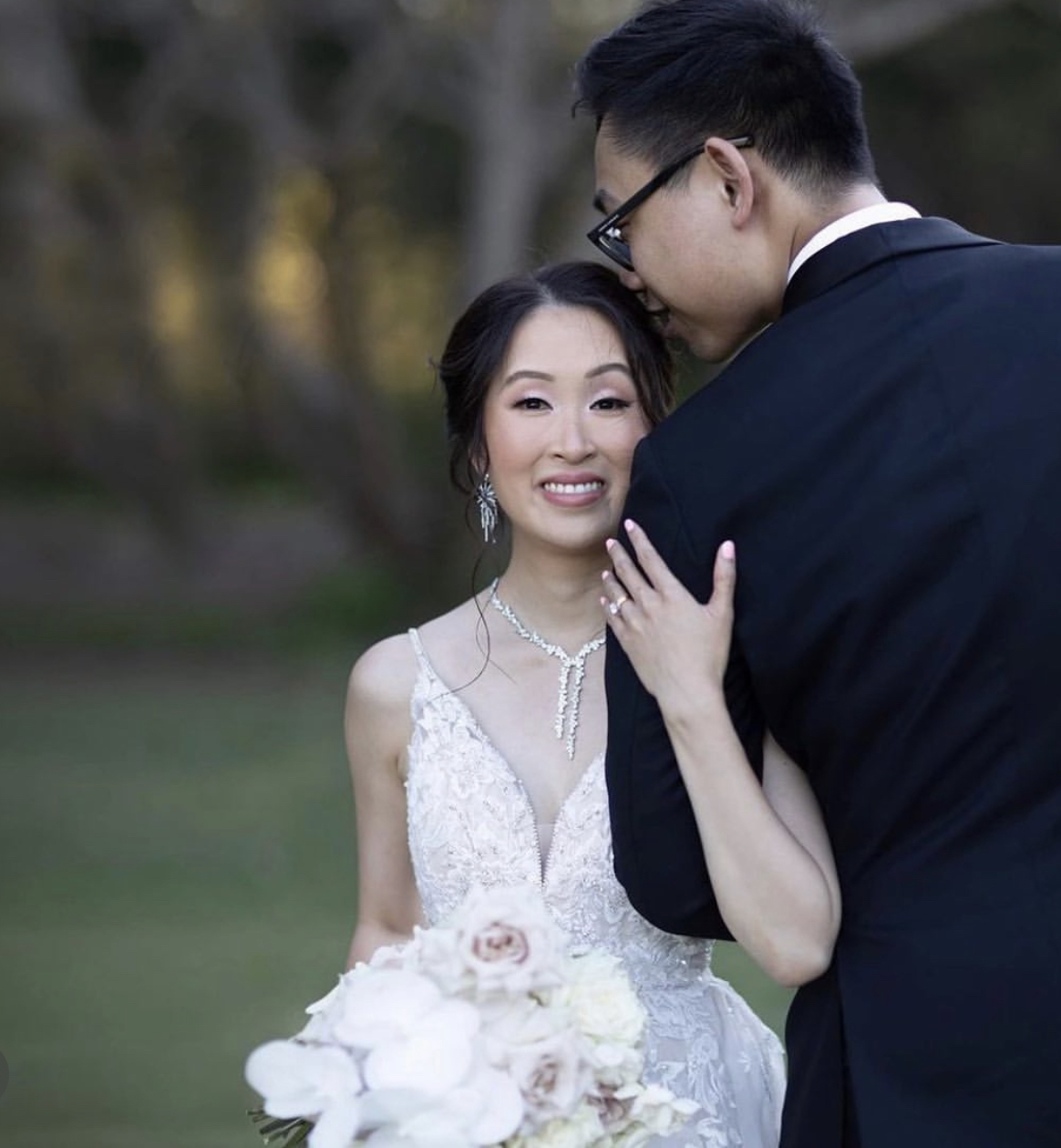 A bride in a white wedding dress and a groom in a black suit share an intimate moment outdoors, with the groom leaning his forehead against the bride's head, and she smiling gently while holding a bouquet of white and blush flowers.