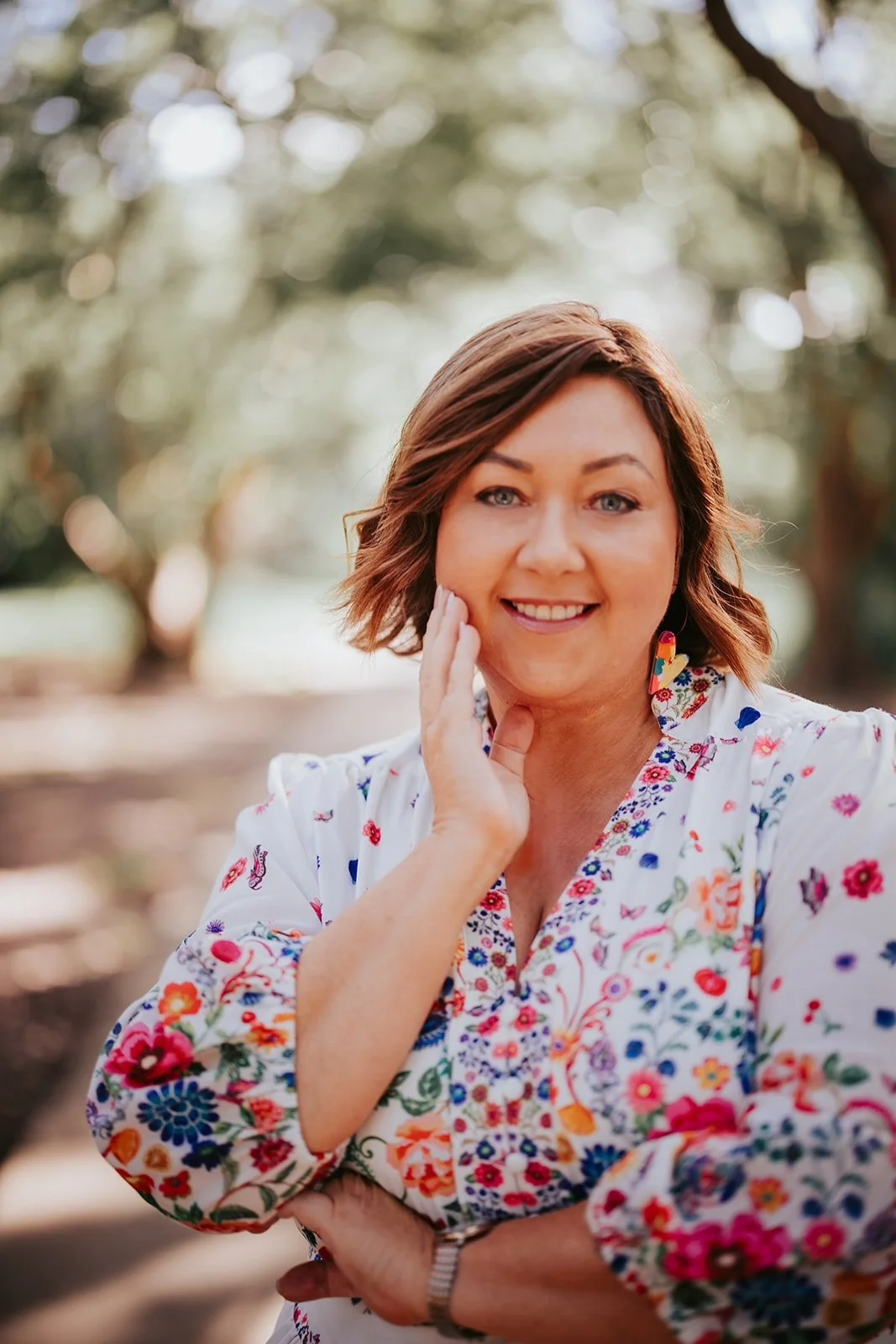 A woman with short brown hair and blue eyes smiling outdoors, wearing a white floral blouse with colorful patterns and earrings, with a blurred natural background.