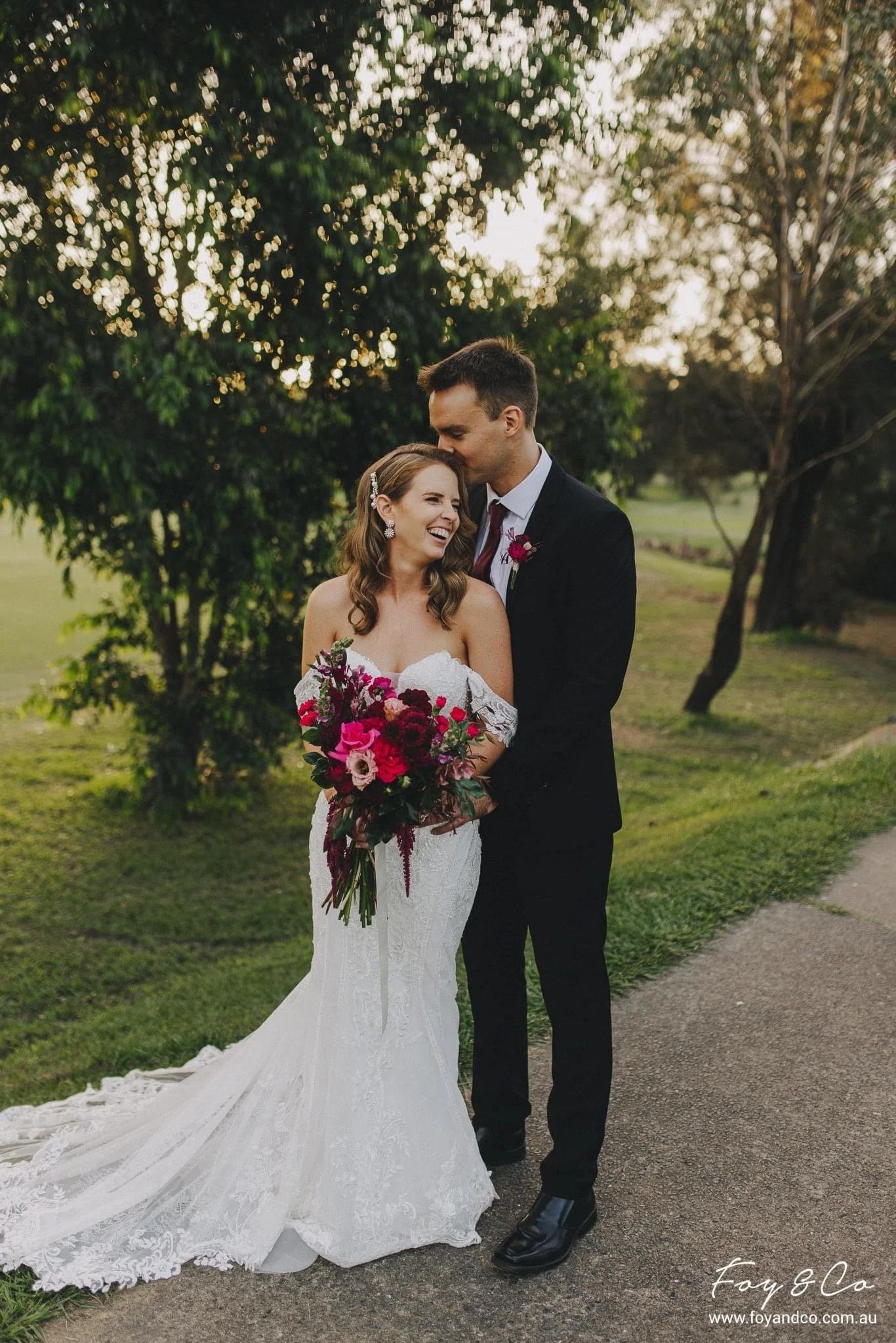 A newlywed couple outdoors, the bride in a white wedding dress holding a bouquet of red, pink, and purple flowers, smiling and laughing, the groom in a black suit with a pink boutonniere, kissing her on the forehead, surrounded by trees and greenery 