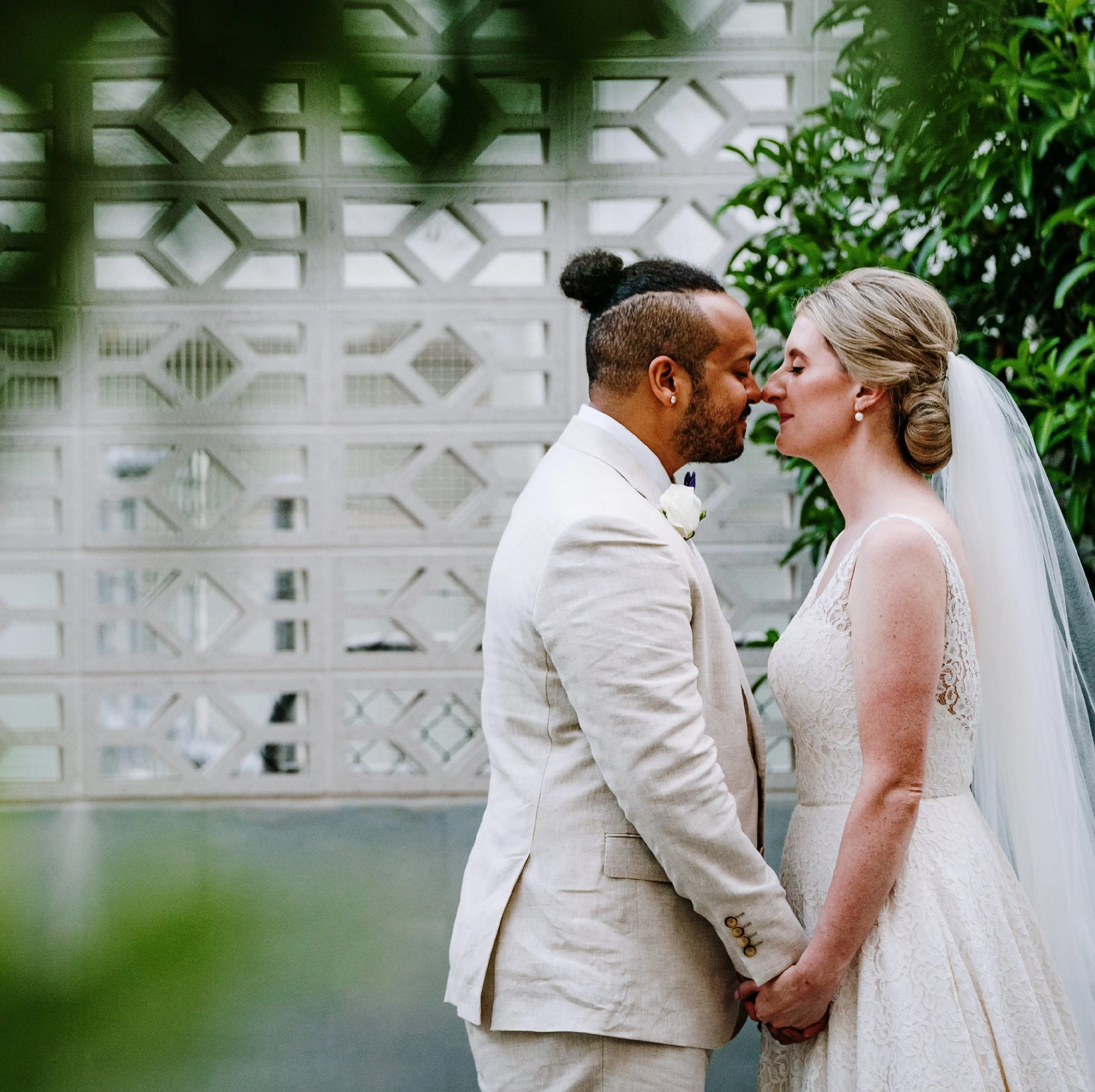 A couple on their wedding day, standing closely with foreheads touching and holding hands. The groom has dark skin and a hairstyle with a bun, wearing a light-colored suit. The bride has fair skin, blonde hair in an elegant updo, wearing a lace weddi