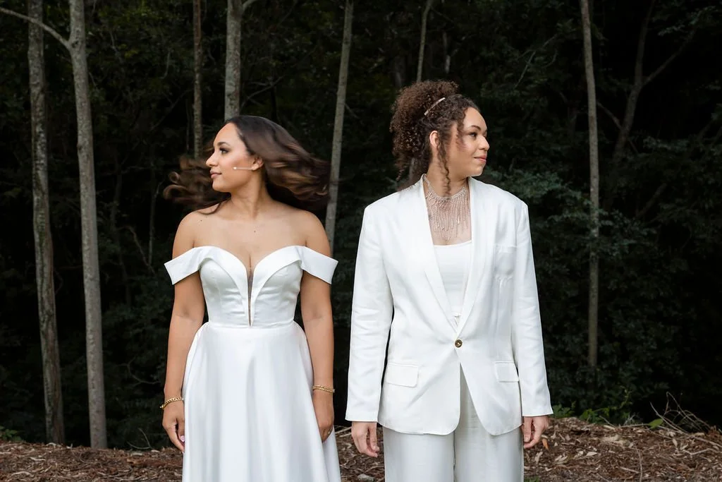 Two women standing outdoors in a wooded area, one in a white off-shoulder wedding dress with flowing hair, and the other in a white suit with curly hair, both looking to the side.