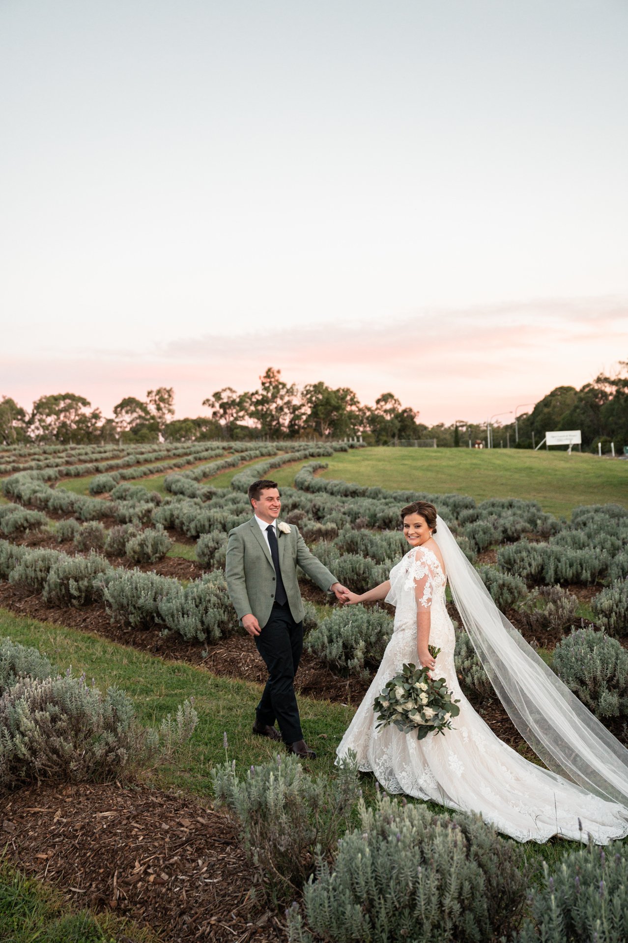 A bride and groom holding hands and smiling in a lavender field at sunset. The bride wears a white wedding gown with a long train and veil, holding a bouquet of greenery. The groom wears a gray blazer, white shirt, and dark tie.