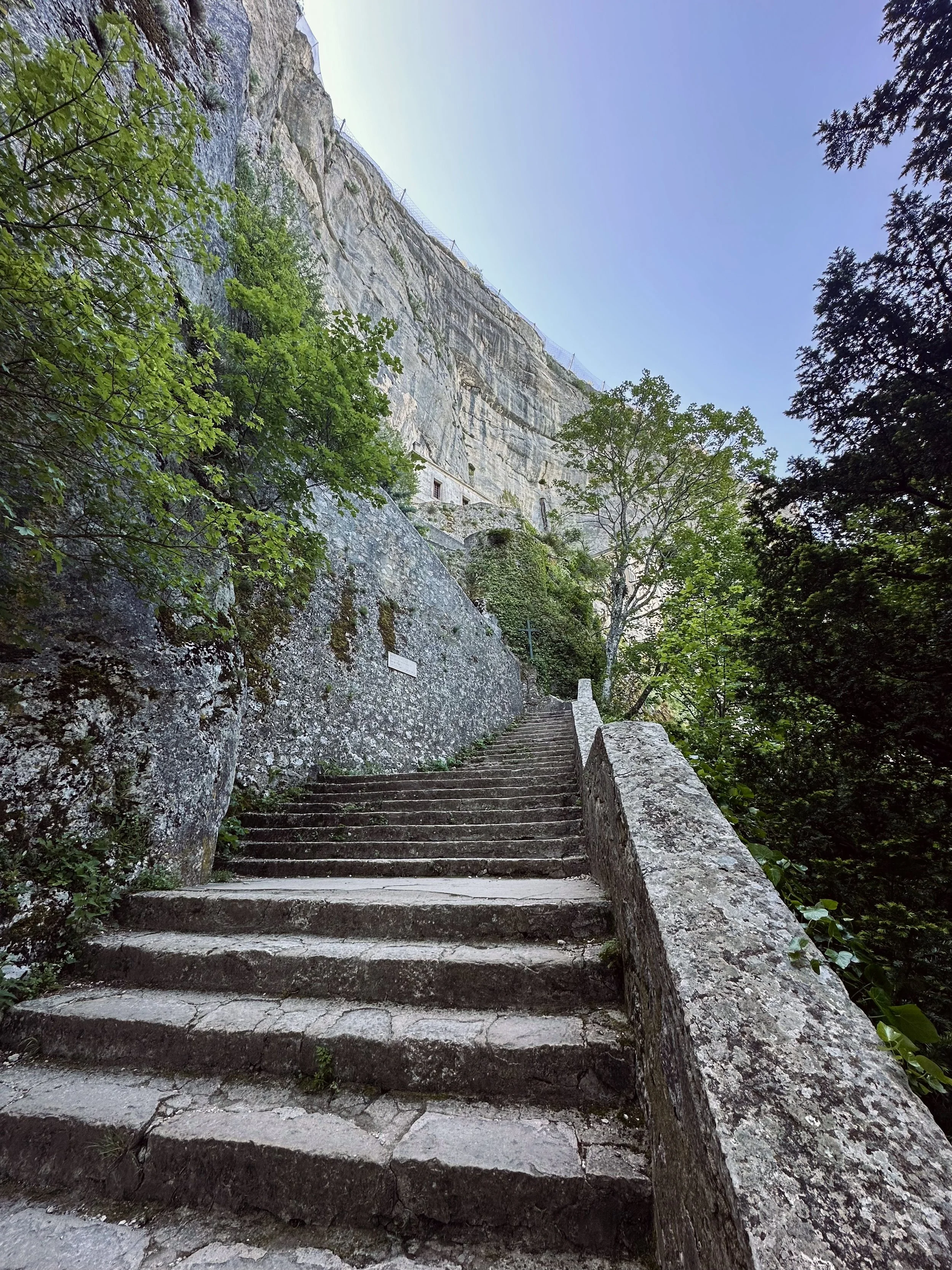 Stone staircase ascending along a rocky mountainside with trees and greenery on either side and buildings carved into the cliff