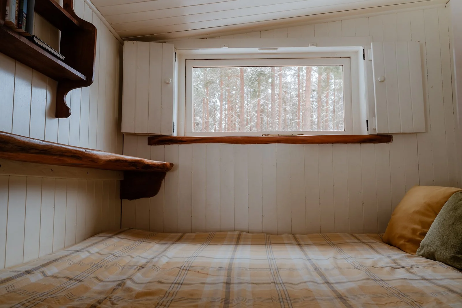 A cozy bedroom with white wooden panel walls, a window showing a view of a snowy forest, a bed with a plaid blanket, and a couple of pillows. There is a wall corner shelf on the left side of the window.