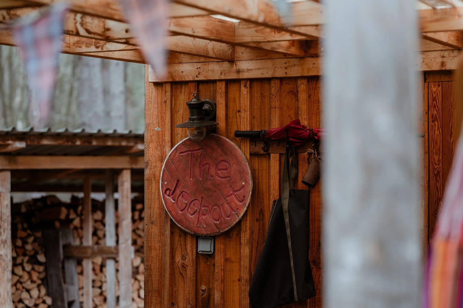 A wooden shed with a round sign reading 'The dock all' and a black lantern. Hanging on the shed's wall are a red umbrella and black bag, with a background of stacked wood logs.