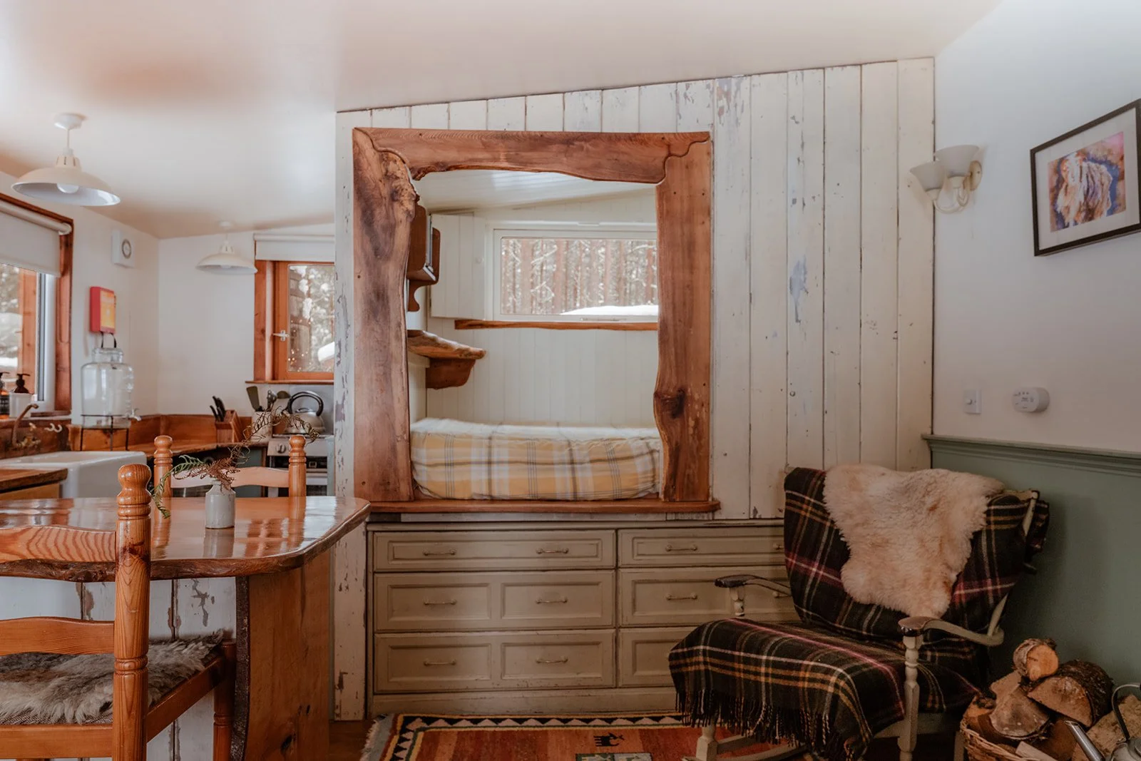 Interior view of a cozy cabin with a sliding wood partition separating a sleeping area with a bed, wood paneling, a window, a plaid chair with a sheepskin throw, and a wooden table.
