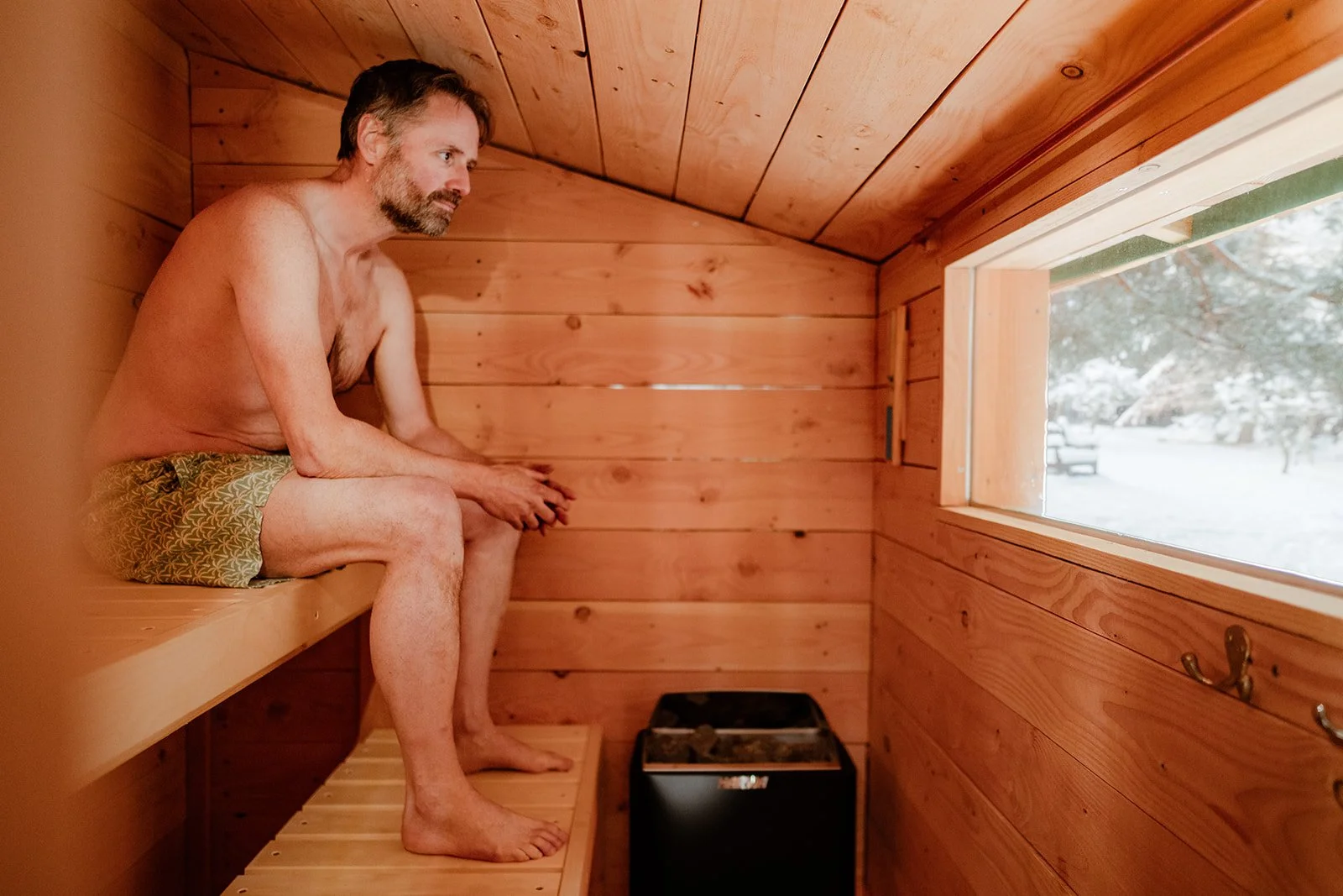 A man sitting alone in a small, wooden sauna, wearing patterned shorts and looking out the window at a snowy outdoor scene.