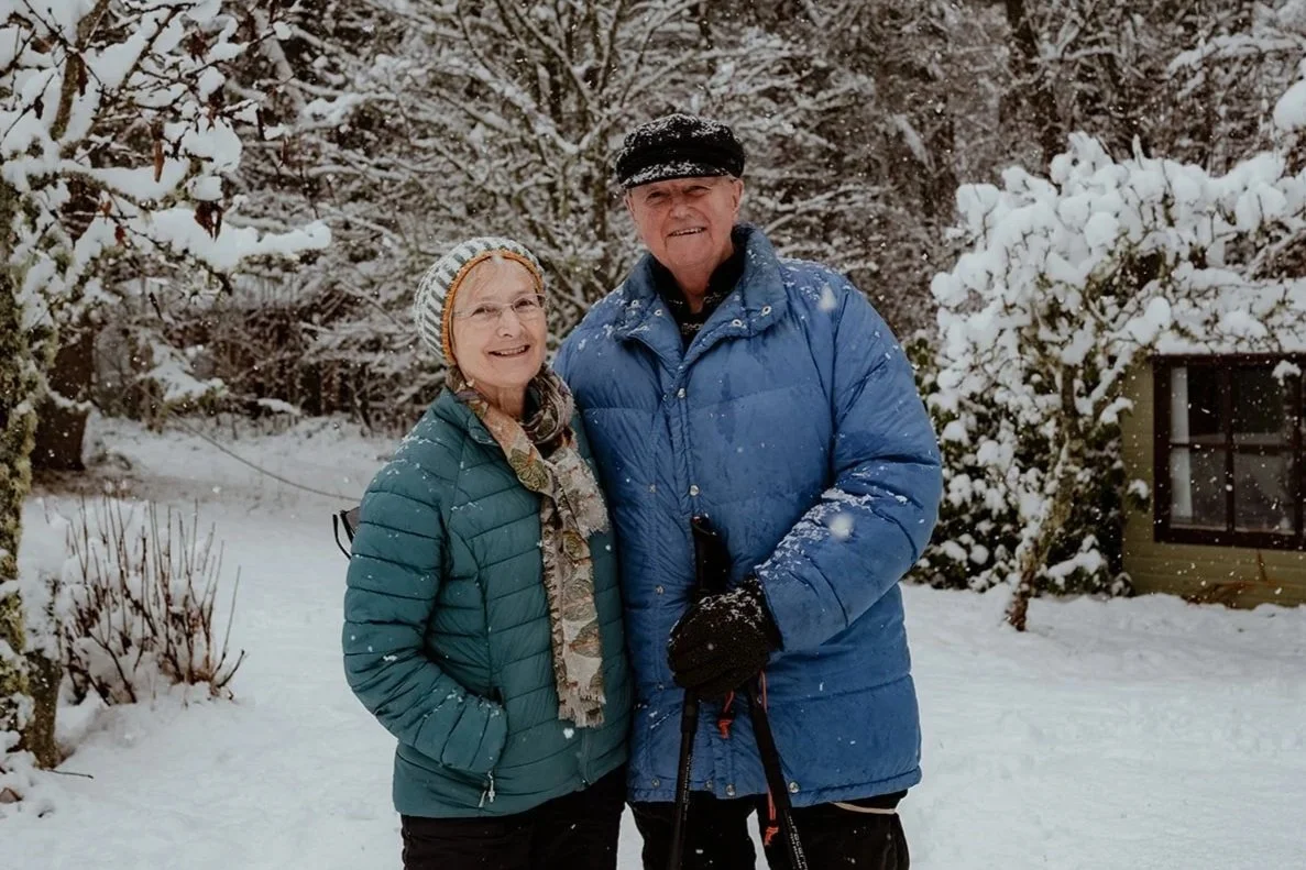 An elderly couple stands outside in snowy weather, smiling, with snow-covered trees in the background. The woman wears glasses, a teal winter jacket, a multicolored scarf, and a striped knit hat. The man wears a blue winter jacket, black gloves, and a black cap, holding walking poles.