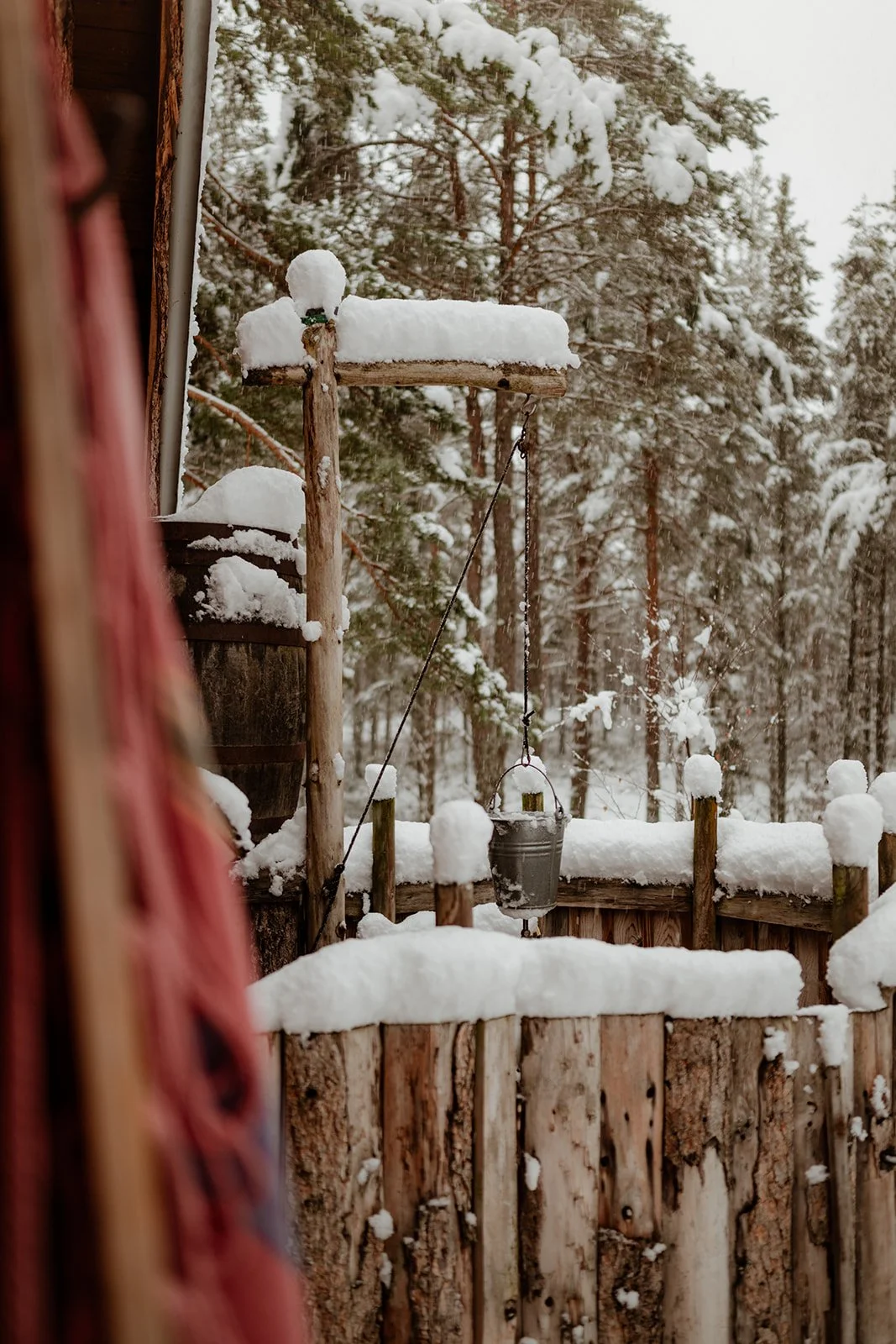 Snow-covered outdoor scene with wooden fences, a hanging bucket, and trees in the background.