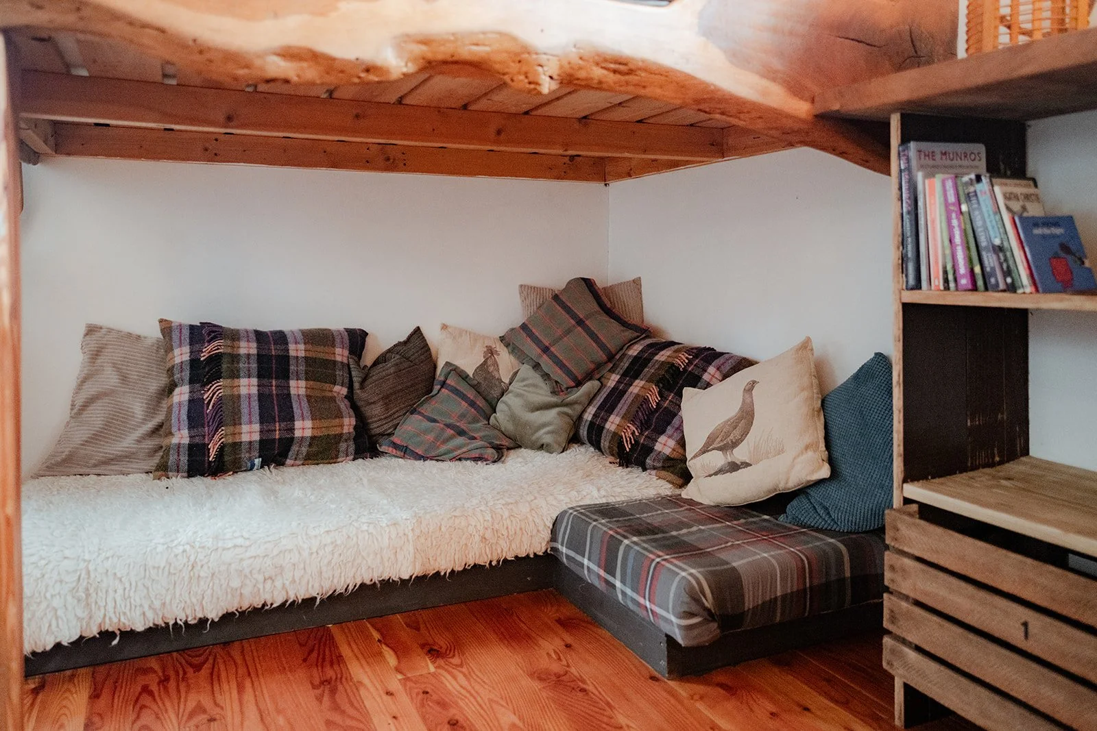 Cozy nook with a low wooden bed frame, layered with plaid and solid throw pillows, covered with a textured white blanket, and flanked by bookshelves filled with books, all set on a wooden floor.